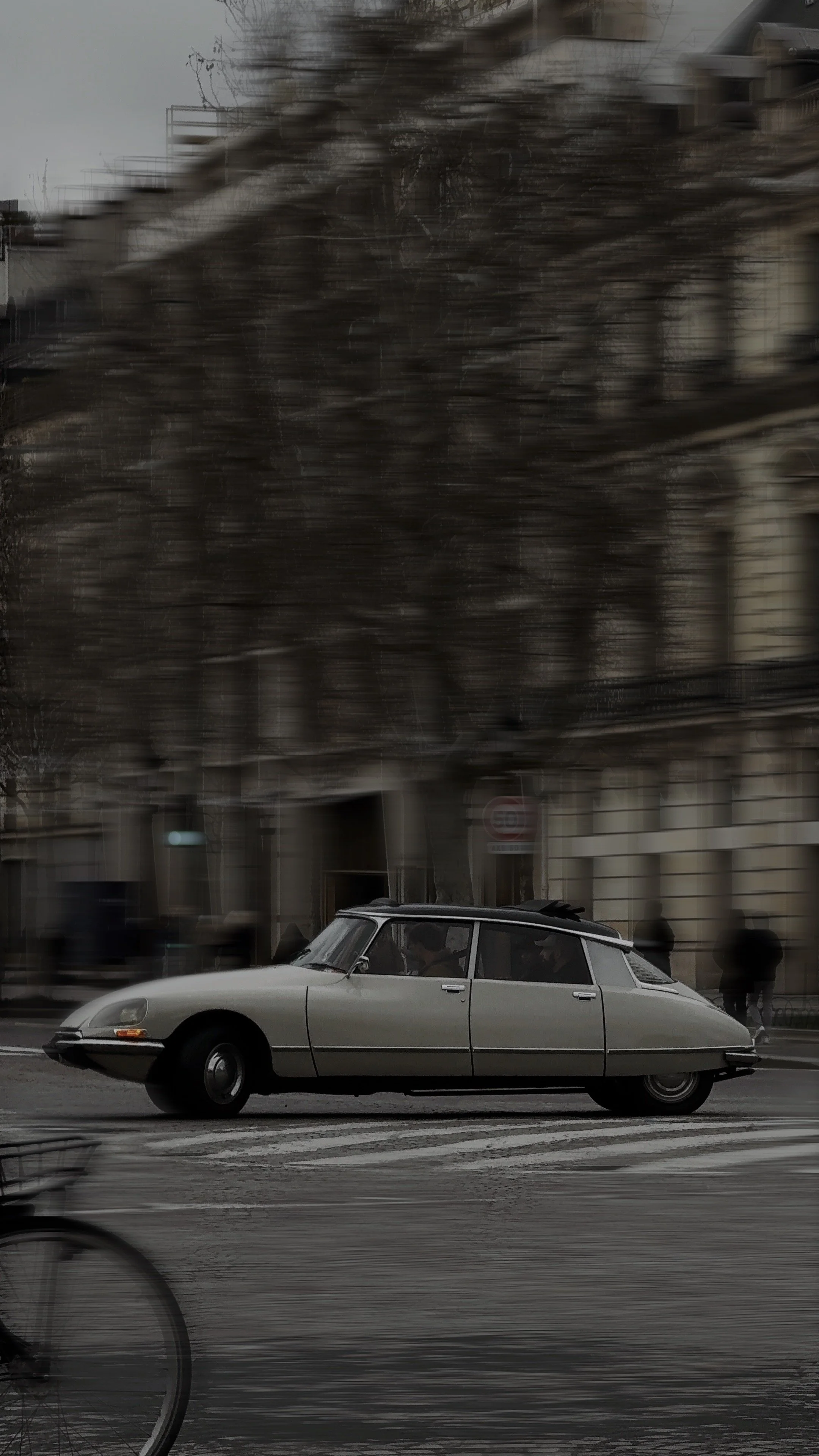 Ein klassischer Citroën DS Wagen fährt auf einer Stadtstraße, im Hintergrund sind Gebäude und Menschen zu erkennen, während im Vordergrund ein Fahrradrahmen sichtbar ist.