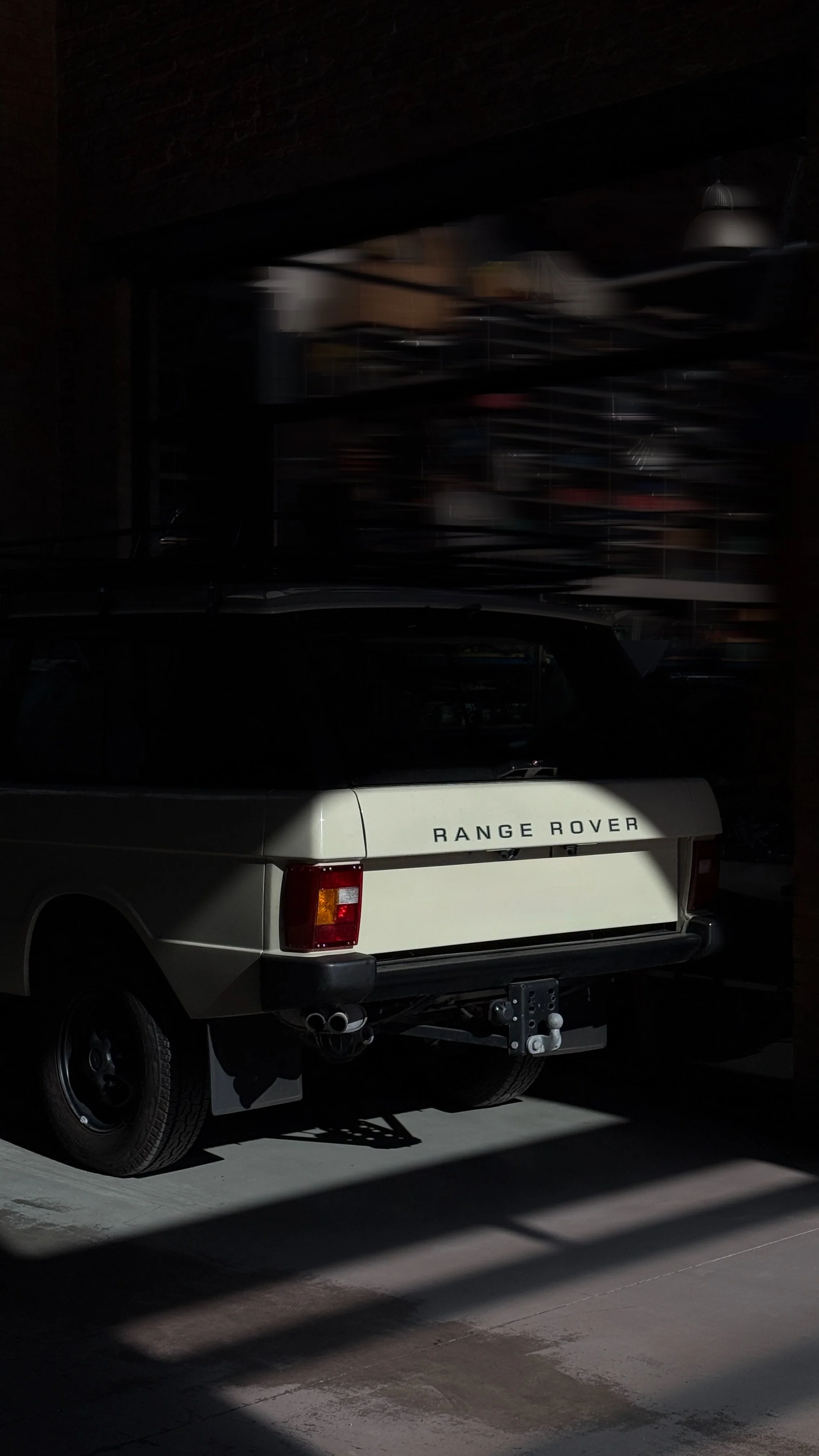 Ein weißer Range Rover in einer Garage, teilweise im Schatten, im Hintergrund ist eine durchsichtige Regalsystem mit Gegenständen. Es ist dunkle Beleuchtung.