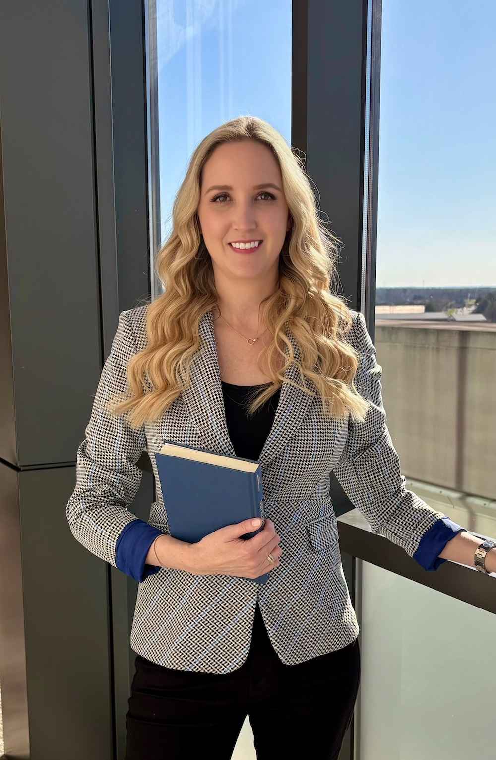 A professional woman with long wavy blonde hair standing by a window, smiling, holding a blue hardcover book, wearing a houndstooth blazer with blue cuffs, black top, and black pants.