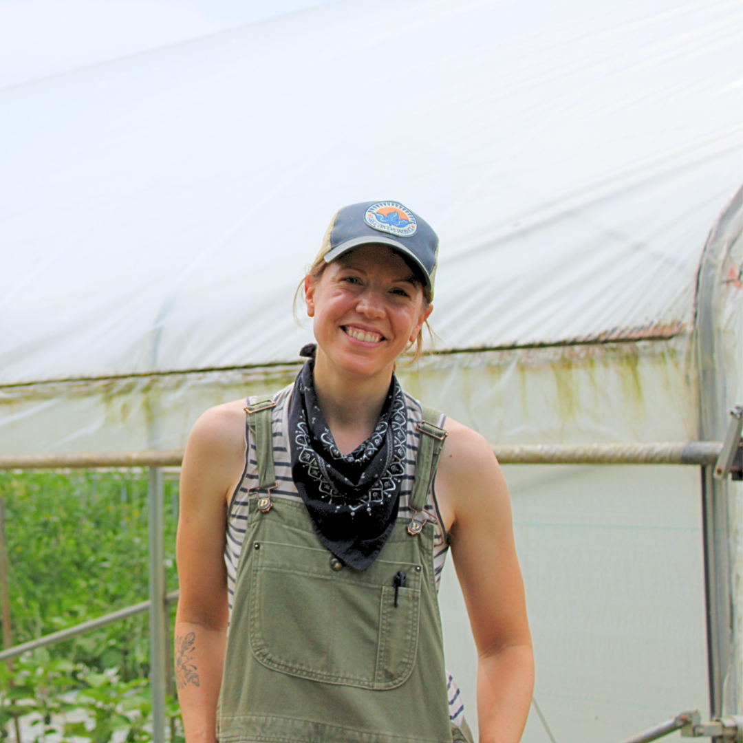 A woman with red hair and a black bandana smiling in a greenhouse, wearing a baseball cap, striped shirt, and green overalls.