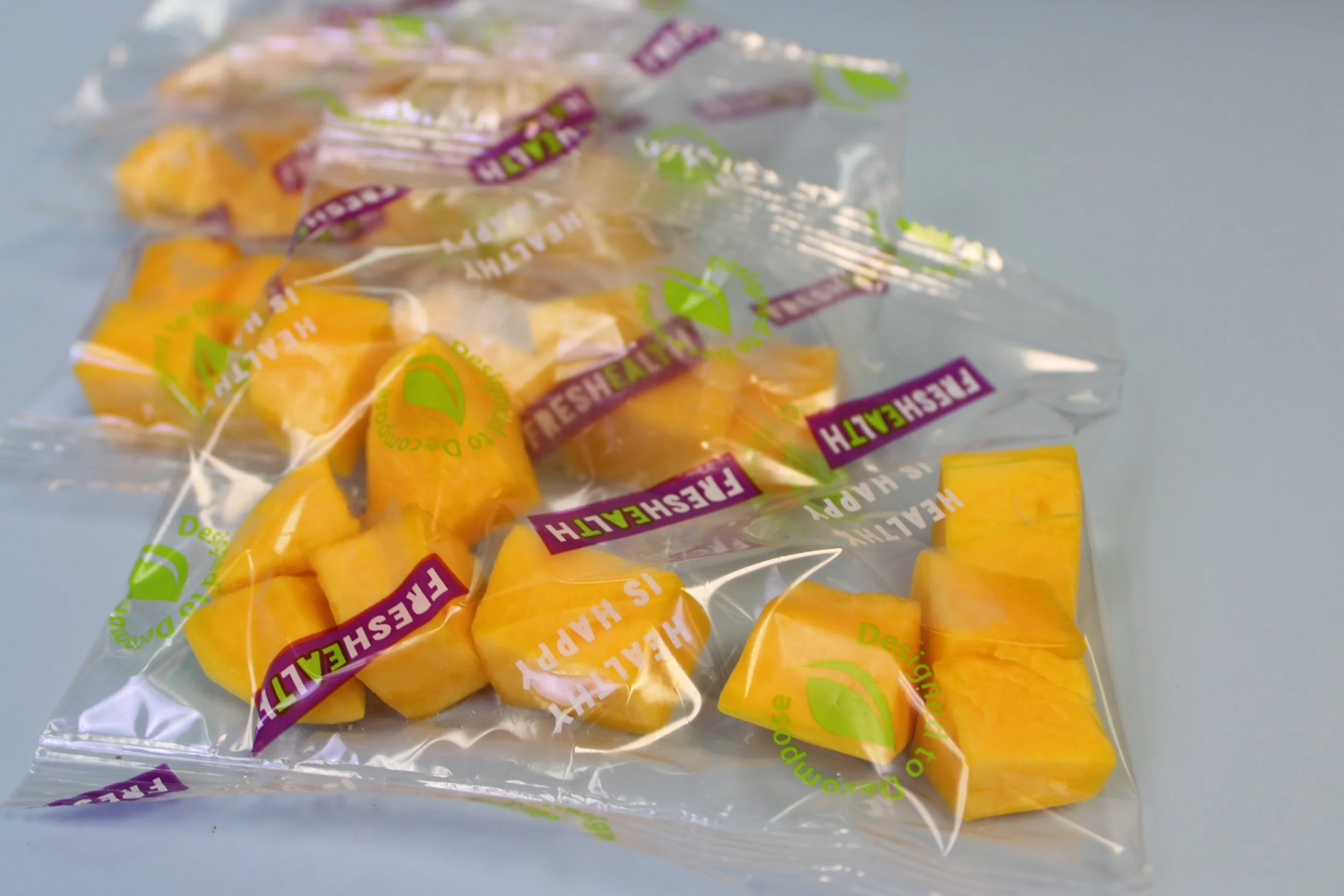 A clear plastic bag with yellow and orange candies inside, branded with green and purple text and logos, resting on a light blue surface.