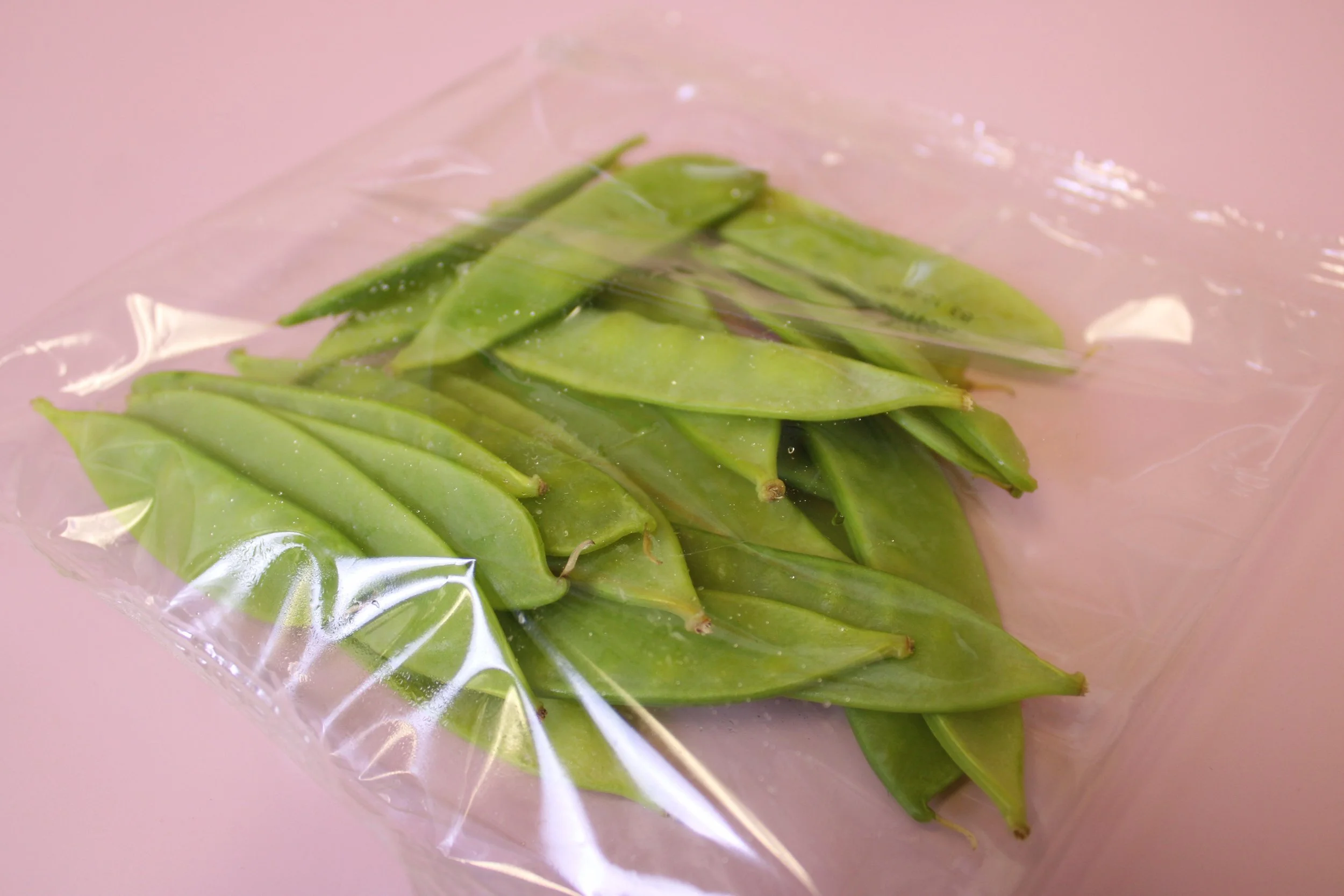 Green snap peas in clear plastic packaging on a pink surface.