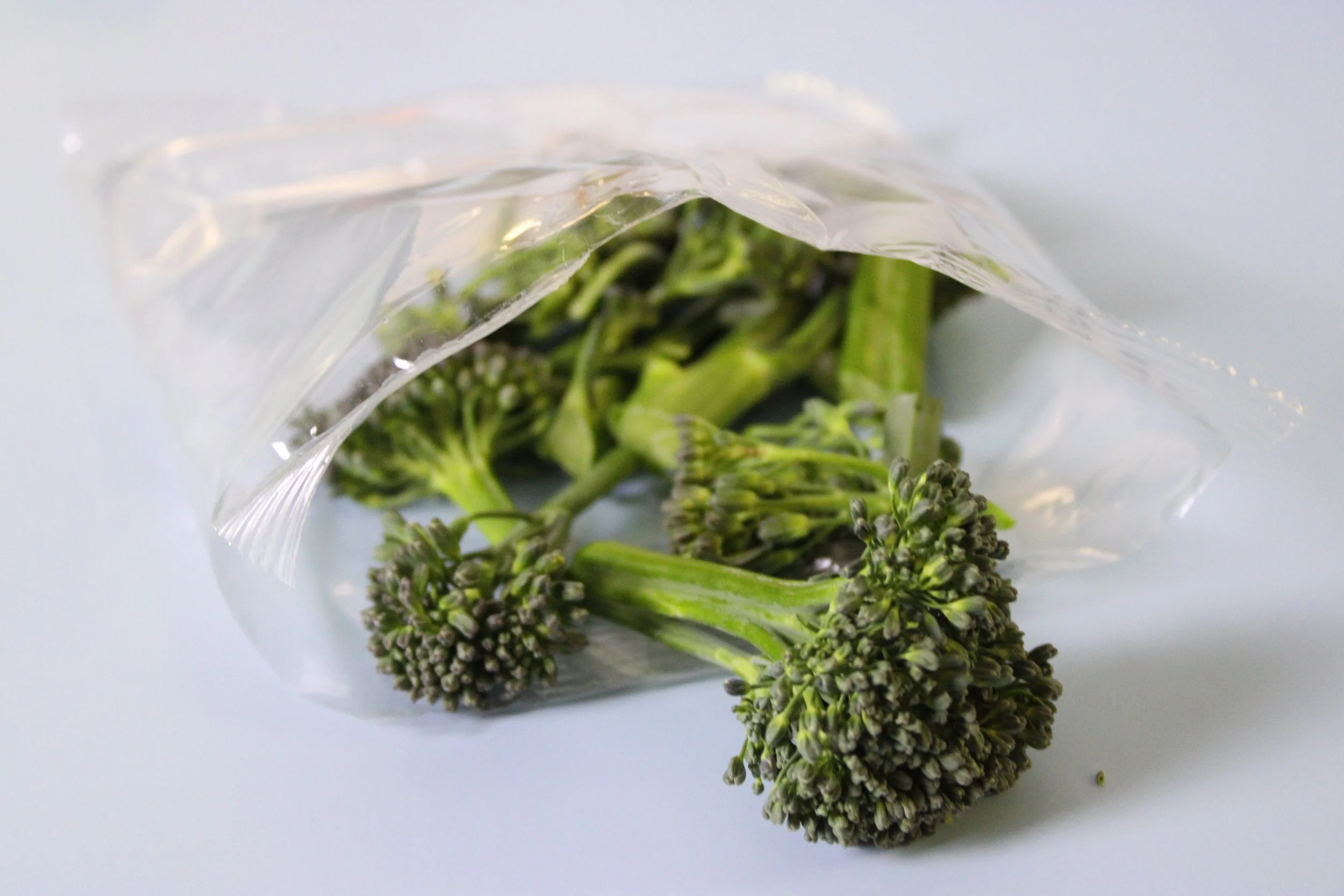 Fresh broccolini in a partially opened plastic bag on a plain white surface.
