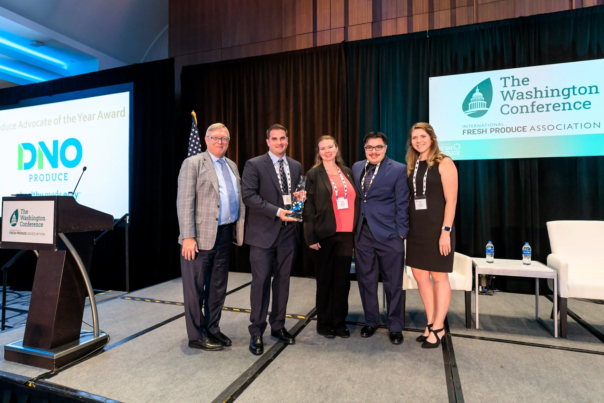 Group of five people on stage at a conference, with a presentation screen showing 'Produce Advocate of the Year Award' and a person holding an award. The background includes a banner for The Washington Conference and the International Fresh Produce Association.