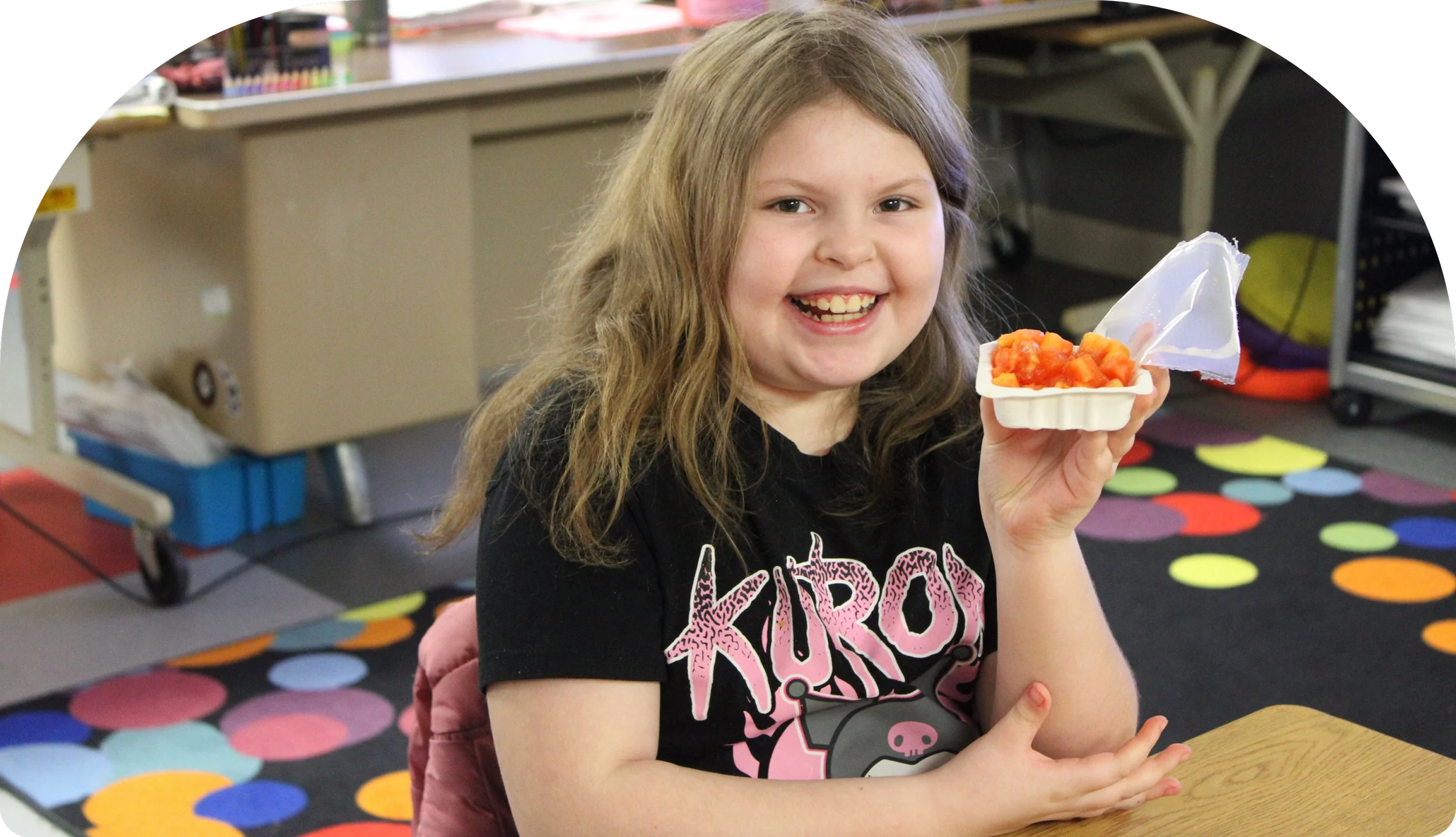 A young girl with long blonde hair, smiling and holding a food container with diced fruit, sitting at a table in a colorful room.