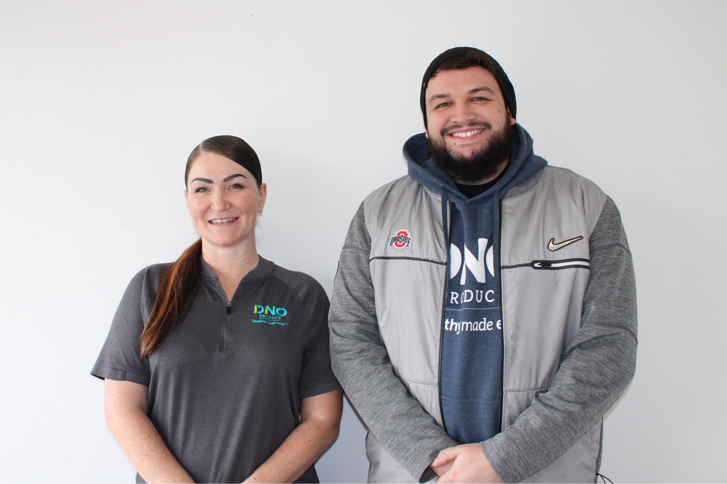 A man and a woman standing side by side against a plain white wall, both smiling at the camera. The woman has long brown hair and is wearing a dark gray athletic shirt with a logo on the chest. The man has short dark hair, a beard, and is wearing a black beanie, a gray and black Nike jacket with patches, and a blue hoodie under the jacket.