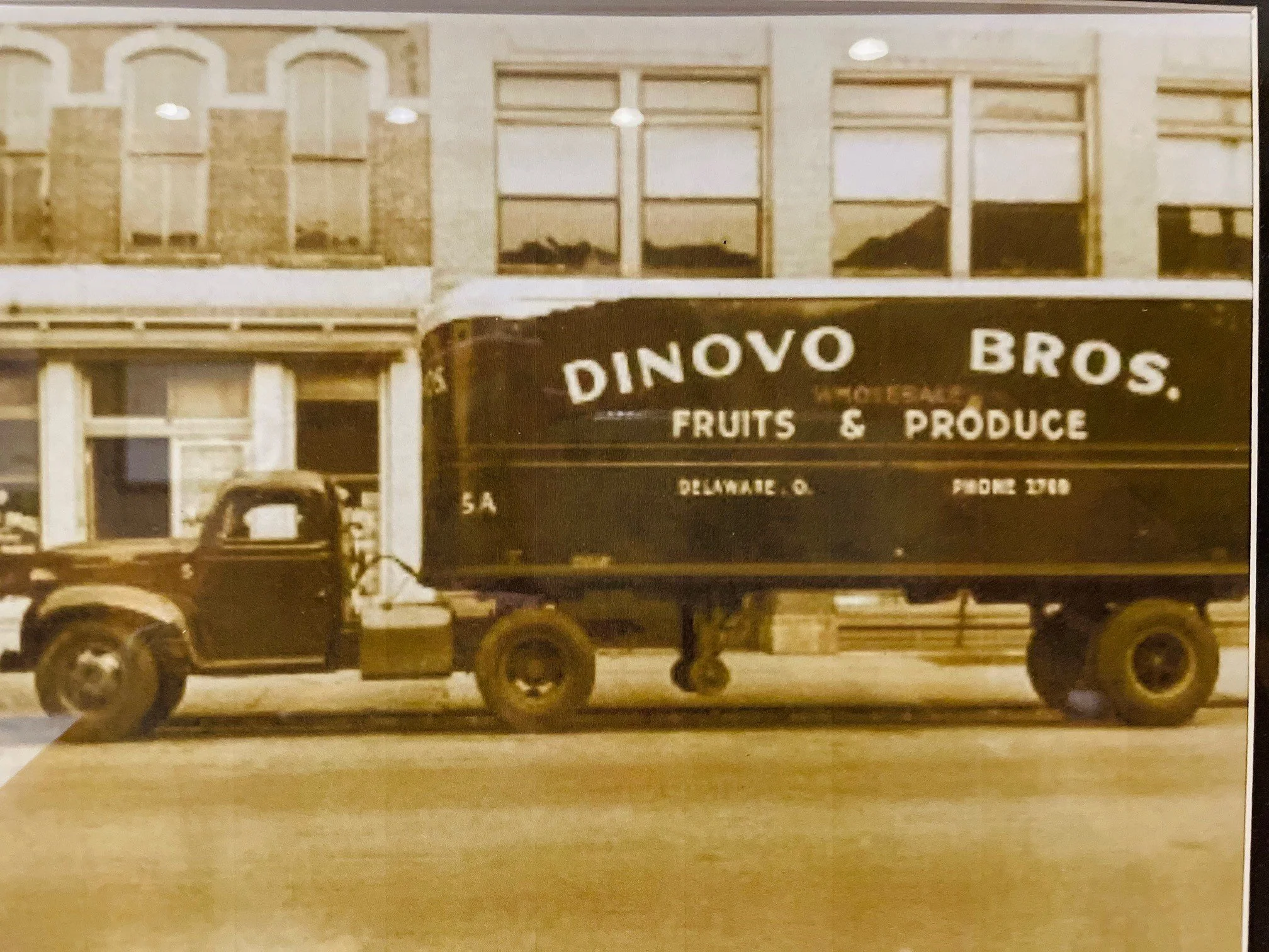 A vintage truck with a black sign that reads 'Dinovo Bros Fruits & Produce' parked in front of a building with multiple large windows.