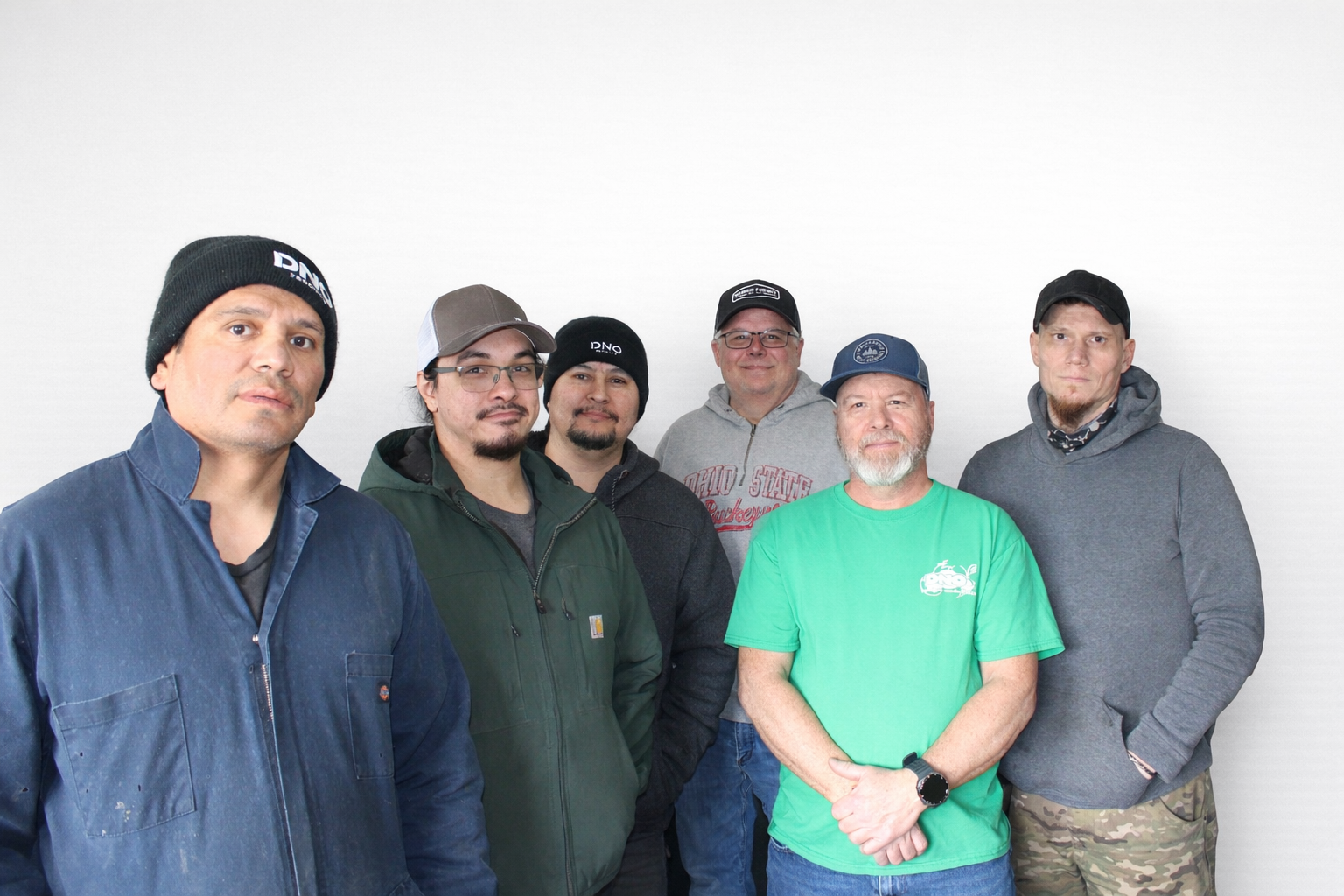 Group of six men standing against a plain white wall, dressed in casual clothing including jackets, hats, and hoodies.