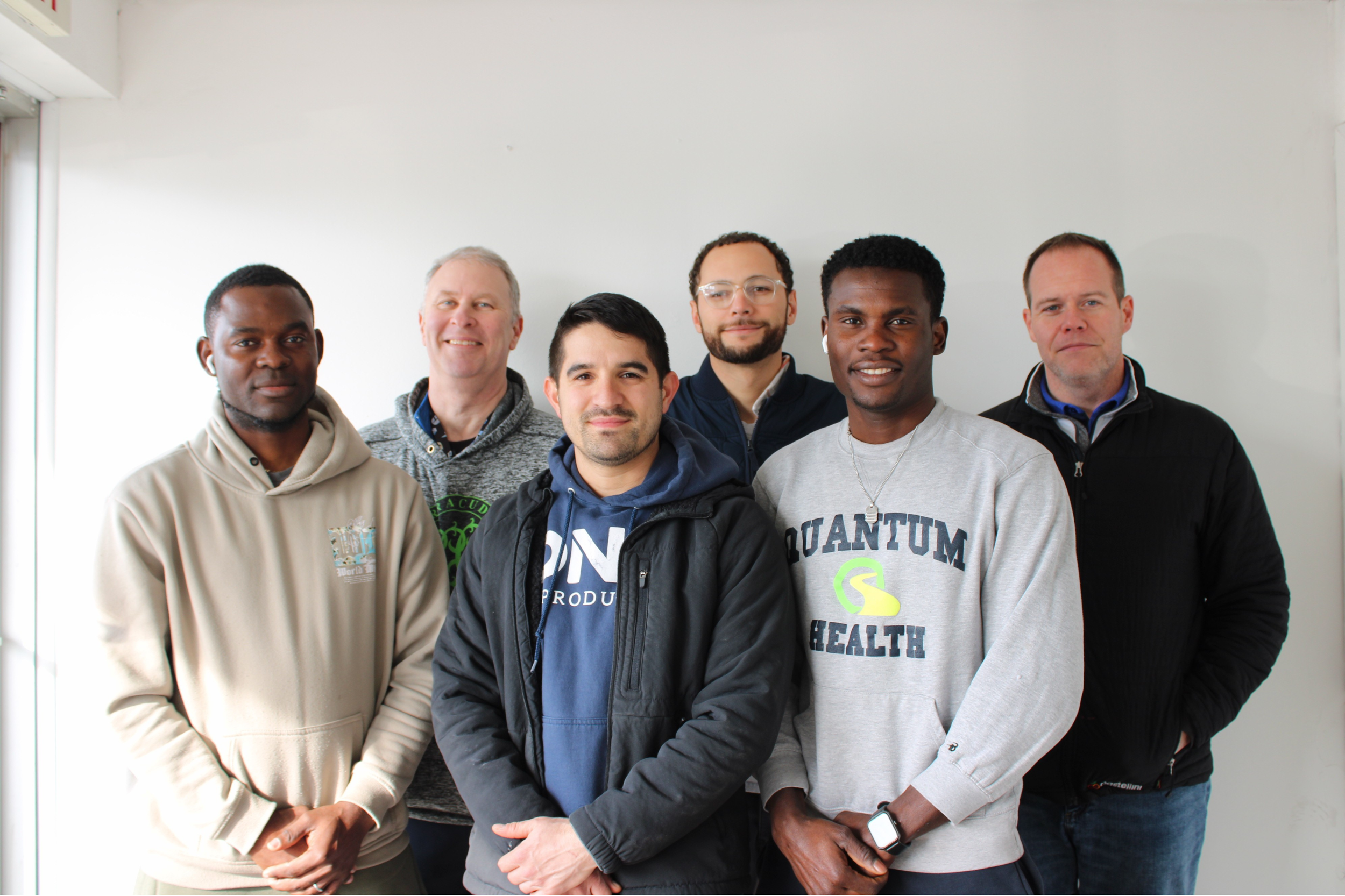 A diverse group of six men standing in a line inside a room with a white wall, some smiling and some with neutral expressions, dressed casually.