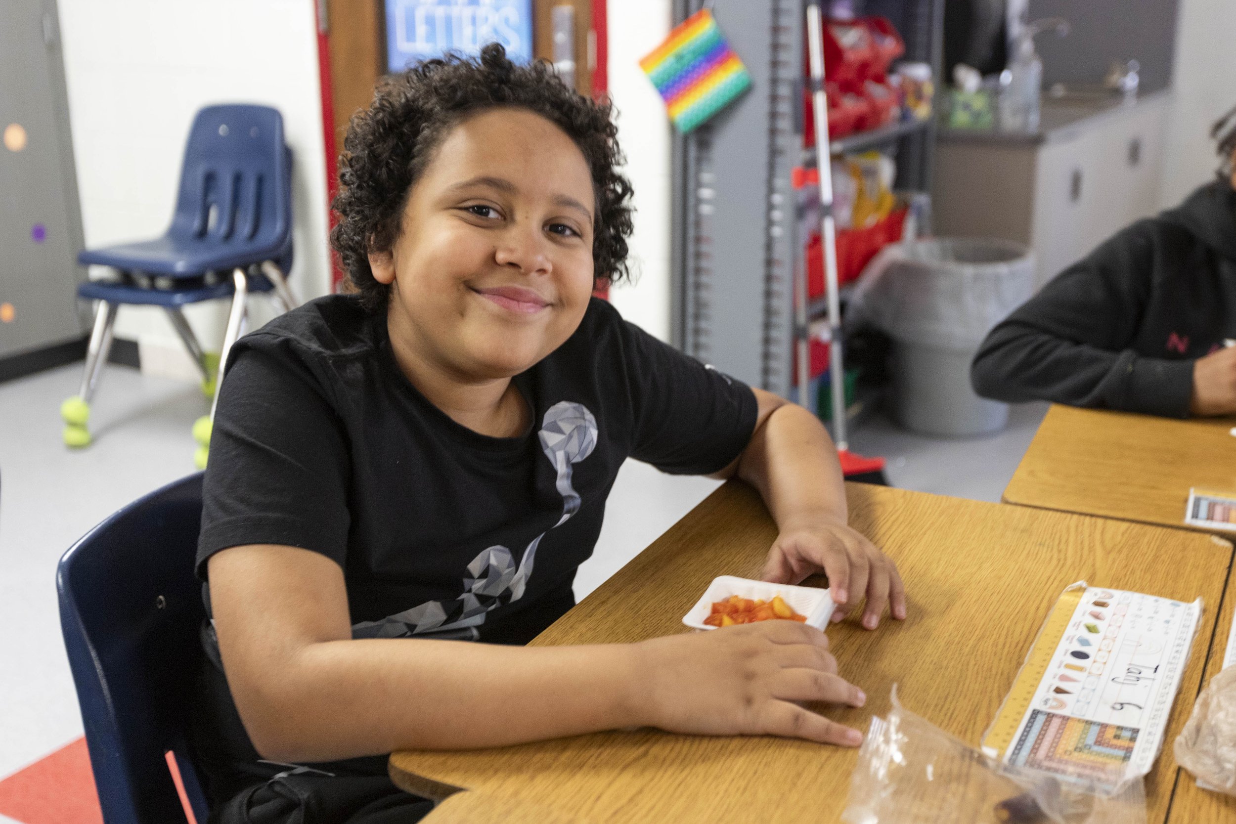 A young boy with curly hair smiling and sitting at a school desk with a tray of orange food, possibly carrots. Behind him are shelves with red containers and a colorful poster, in a classroom setting.