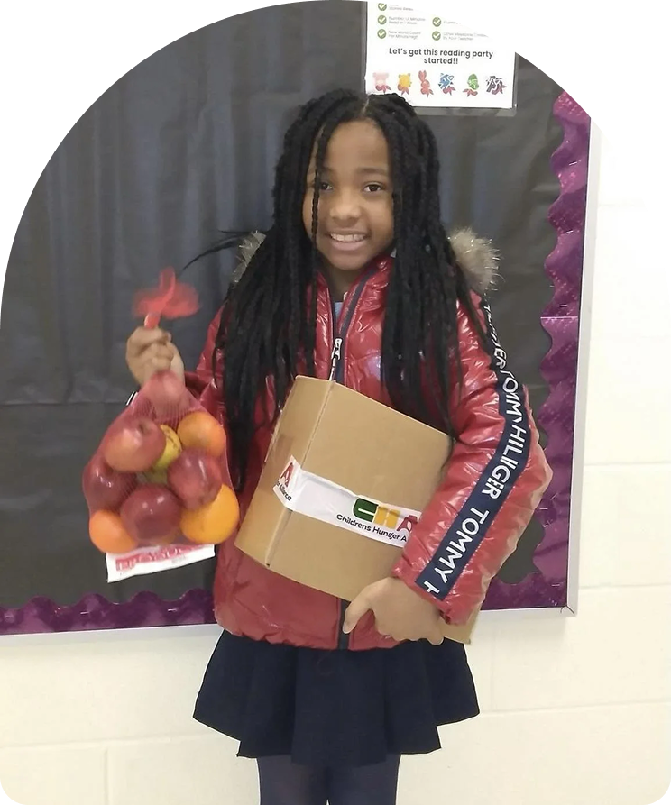 A young girl with long braided hair smiling and holding a bunch of apples and oranges in a plastic bag, along with a cardboard box, standing in front of a bulletin board at school.