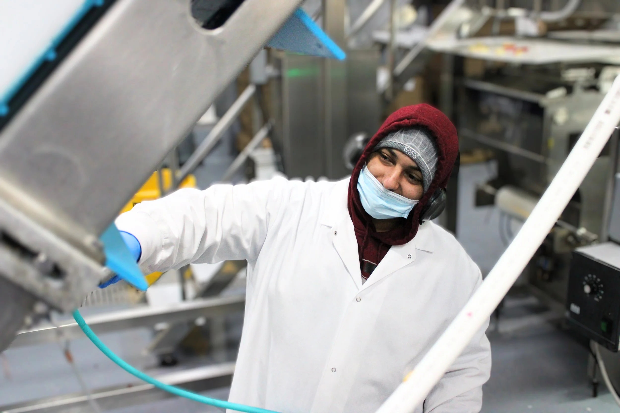 A person in a white lab coat, wearing a gray beanie, a red hoodie, and a face mask, working in a laboratory or industrial setting surrounded by metal equipment.