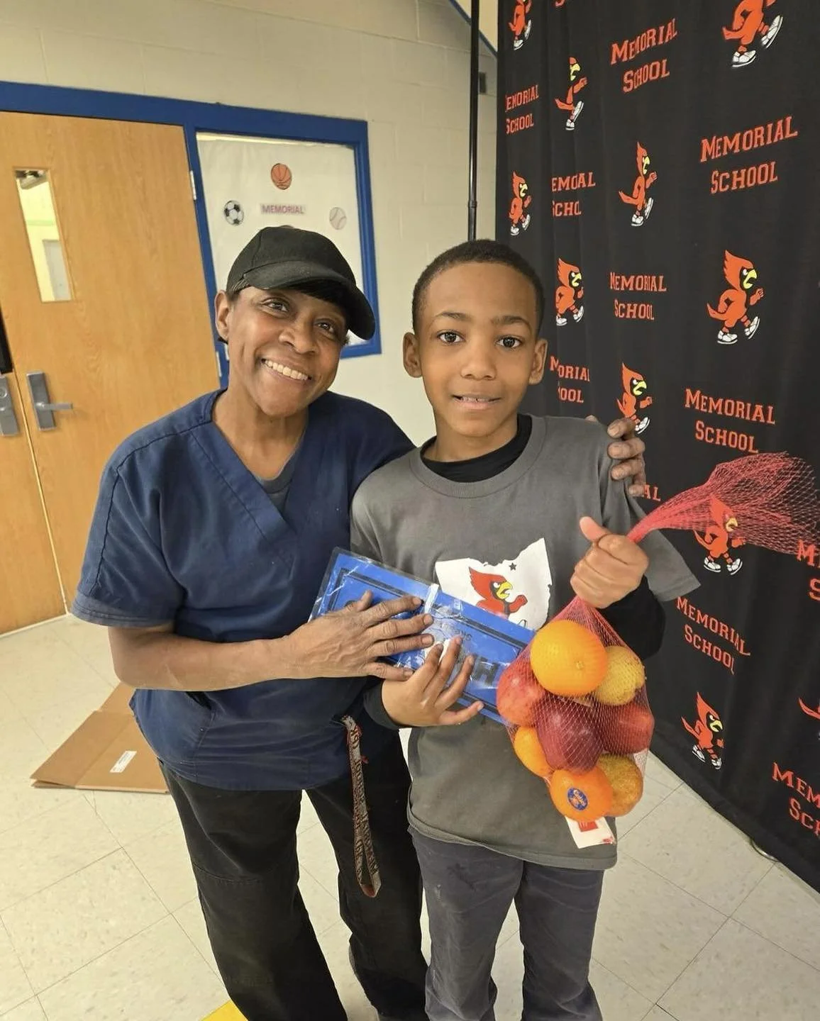 A woman and a boy smiling and posing for a photo at Memorial School. The woman is wearing a navy blue uniform and a black cap, while the boy is holding a net bag with oranges and a squash, and they are standing in front of a backdrop with the school mascot and name.