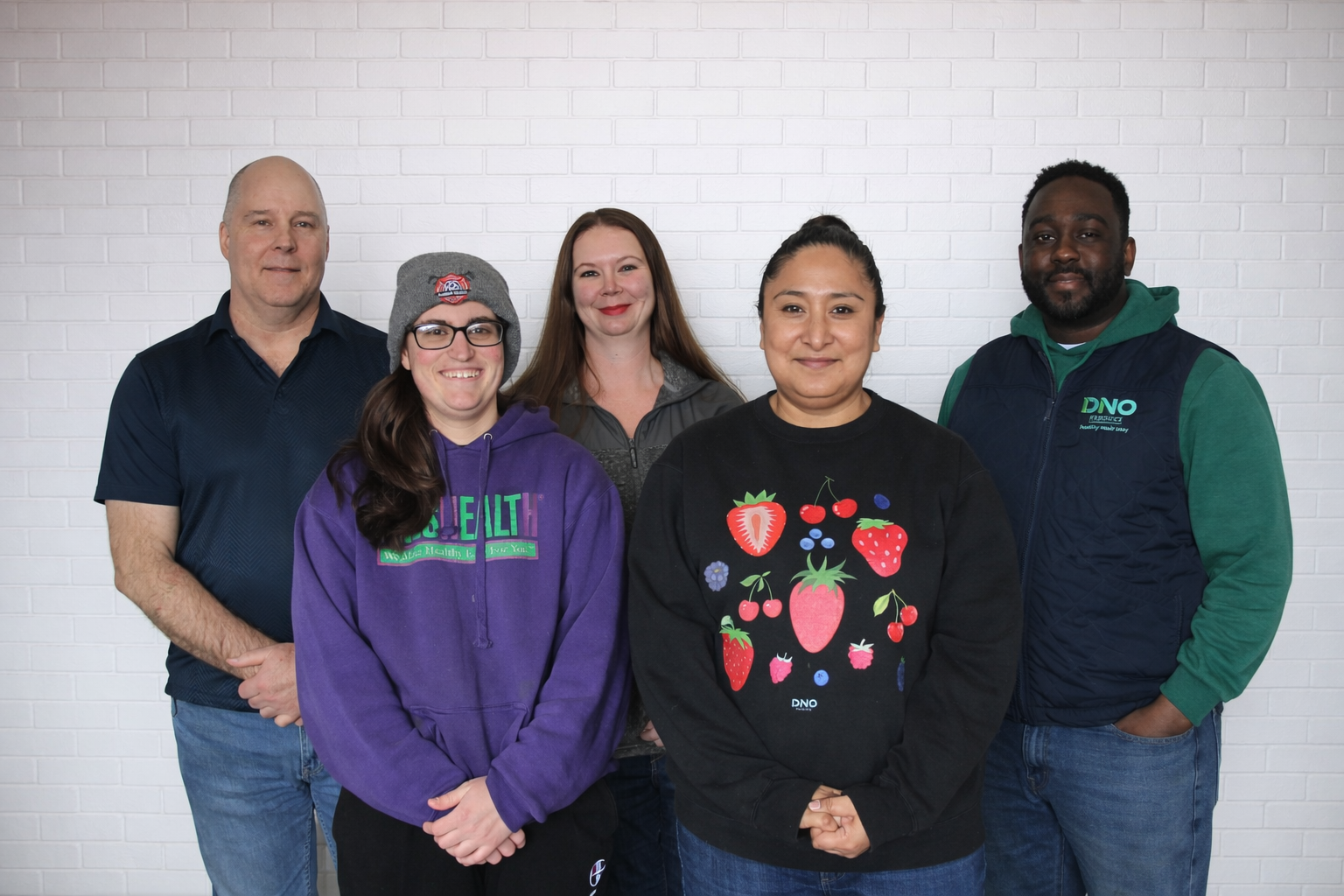 A diverse group of five people standing together in front of a white brick wall, smiling at the camera.