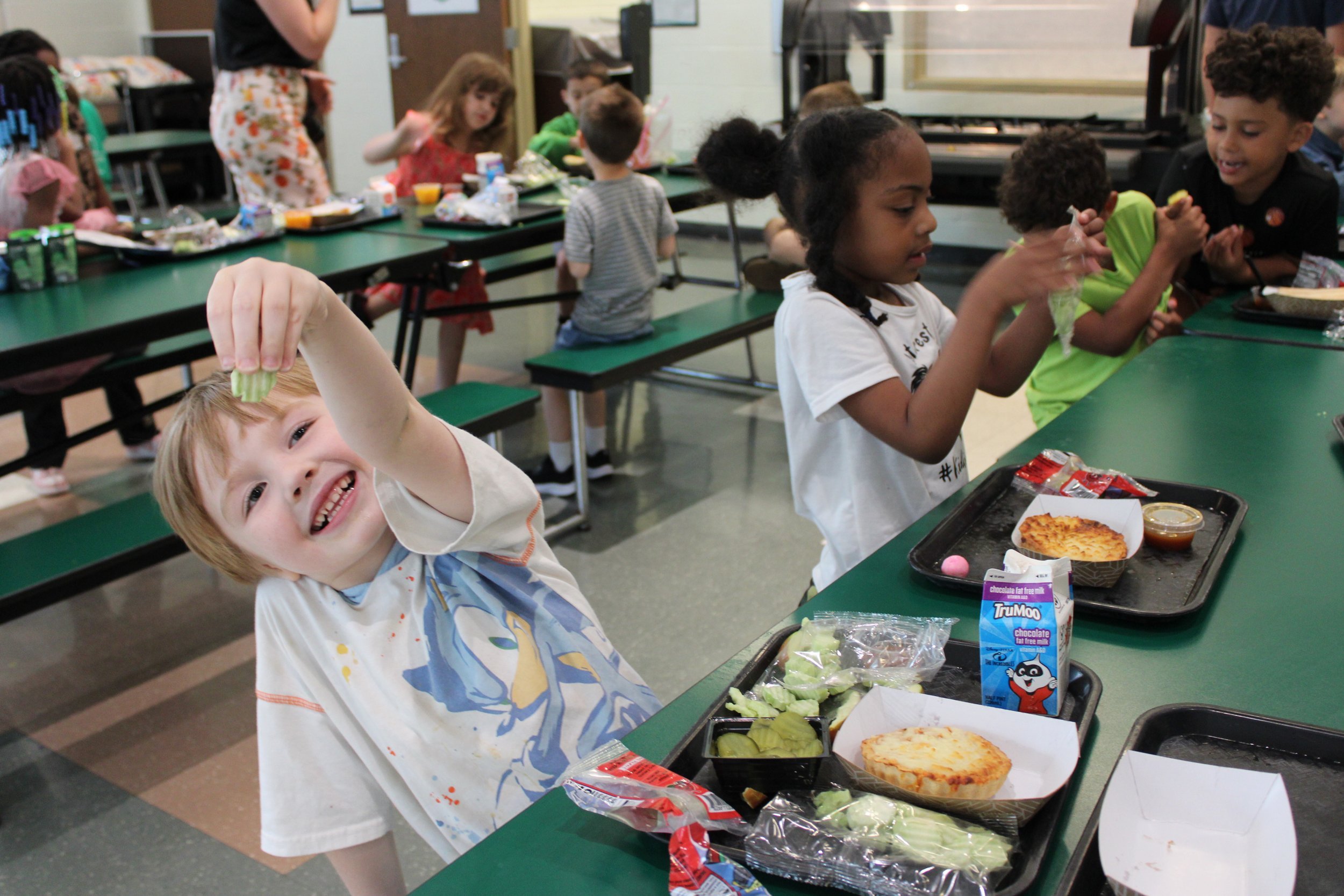 Smiling blonde boy in a children’s hospital gown holding up a celery stalk at a cafeteria table with friends, with trays of food including pizza and a carton of chocolate milk.