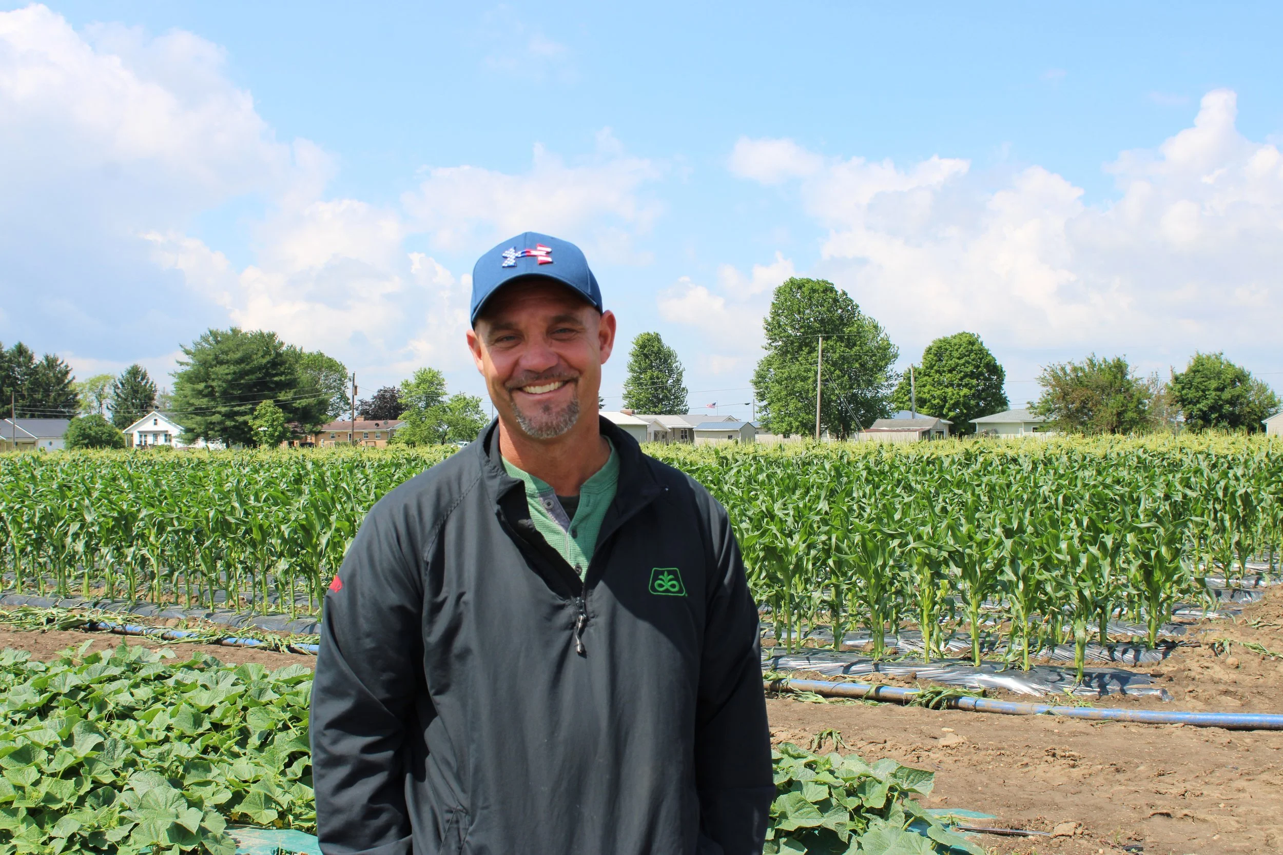 A man smiling in a field of green crops with trees and houses in the background, under a partly cloudy sky.