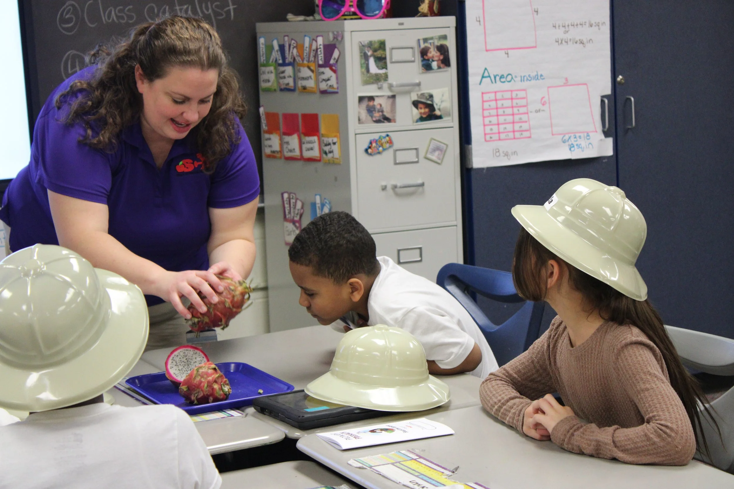 A teacher is demonstrating a fruit, possibly a dragon fruit, to a group of children in a classroom. The children are seated at a table, wearing hats, and looking at the teacher with interest. The classroom has educational posters and pictures on the walls.