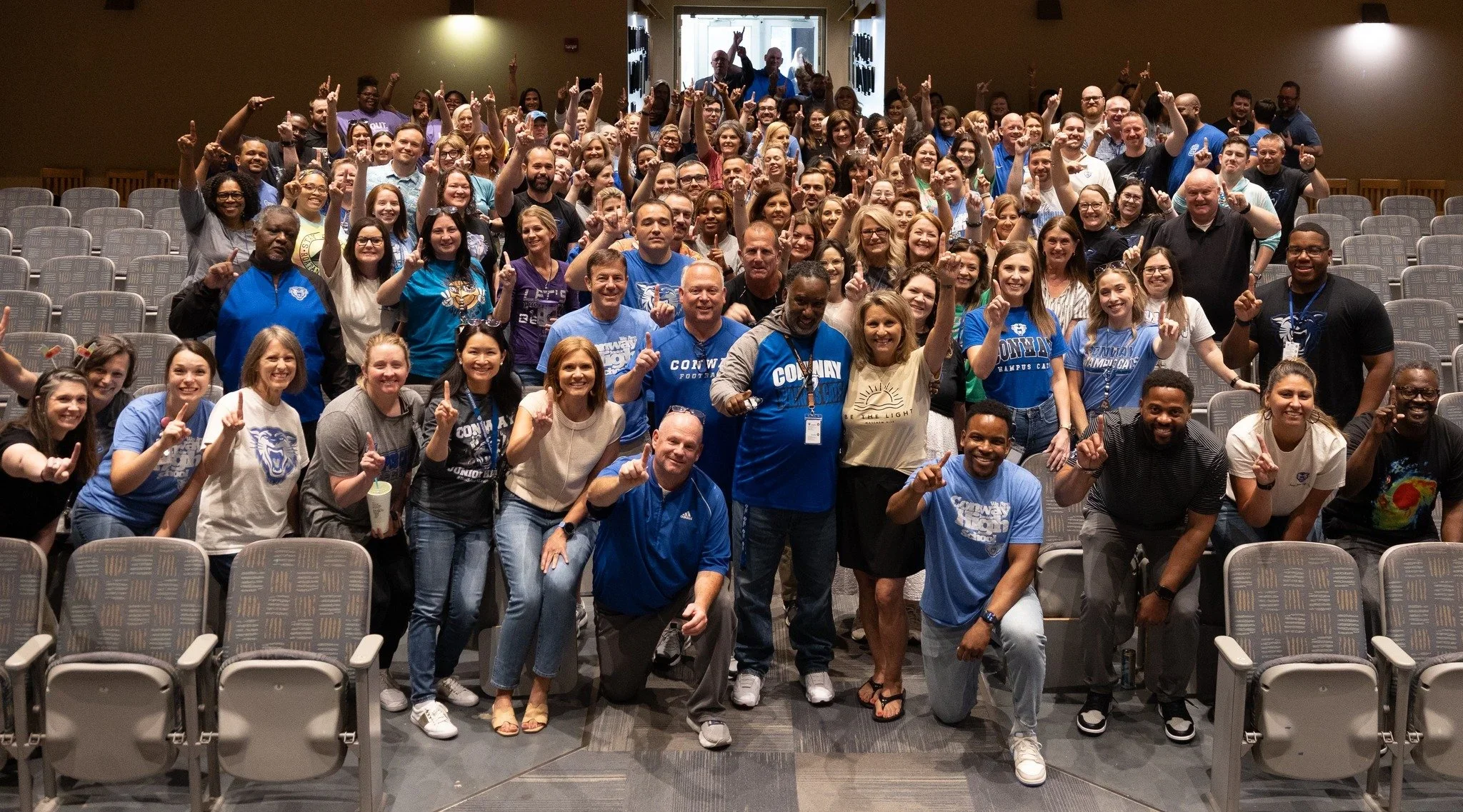 A large group of people gathered in an auditorium, smiling and holding up one finger, with some wearing blue shirts and lanyards, suggesting a team or conference setting.
