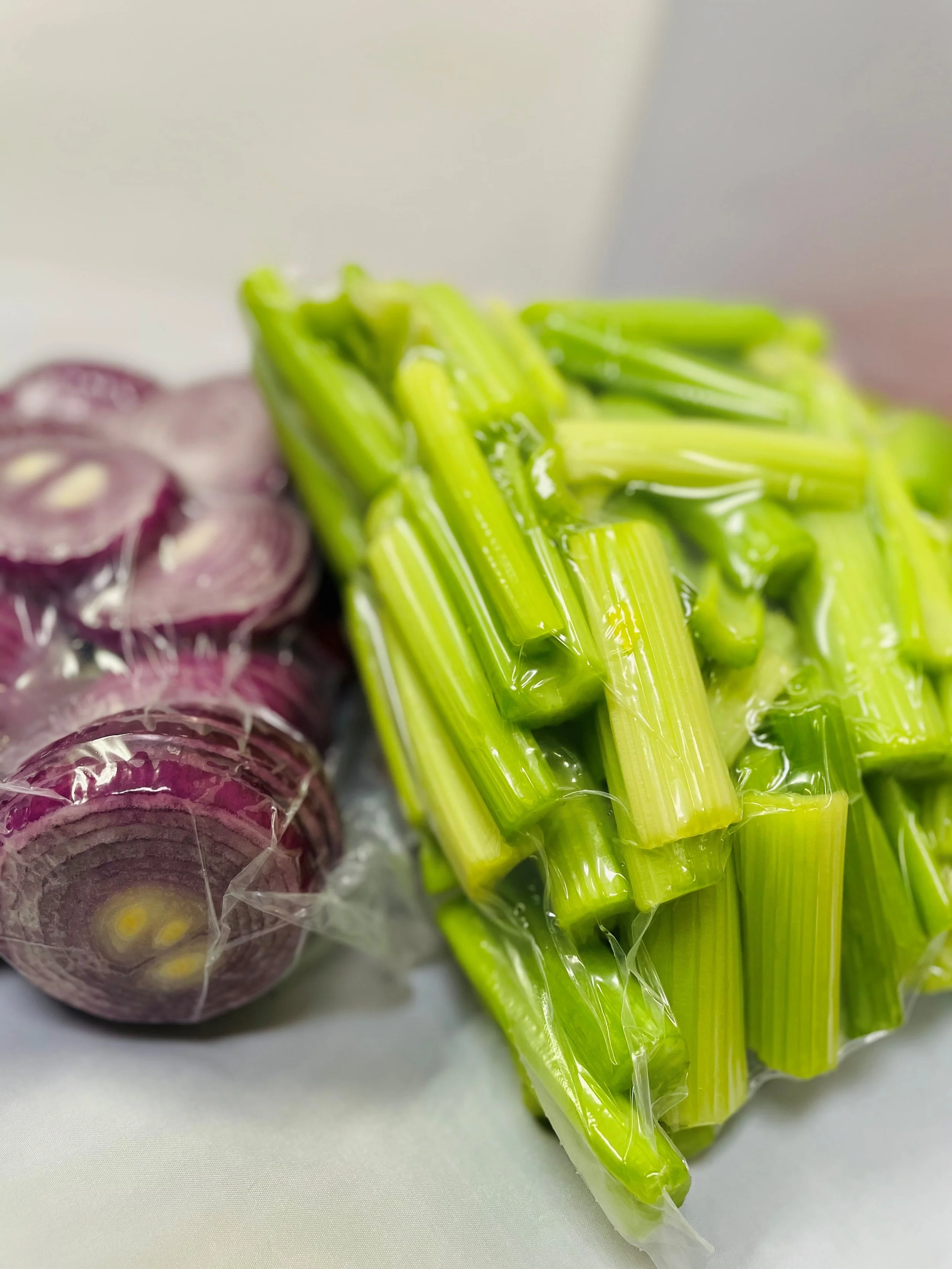 Sliced red onions and a bunch of celery stalks, both in plastic packaging, on a white surface.