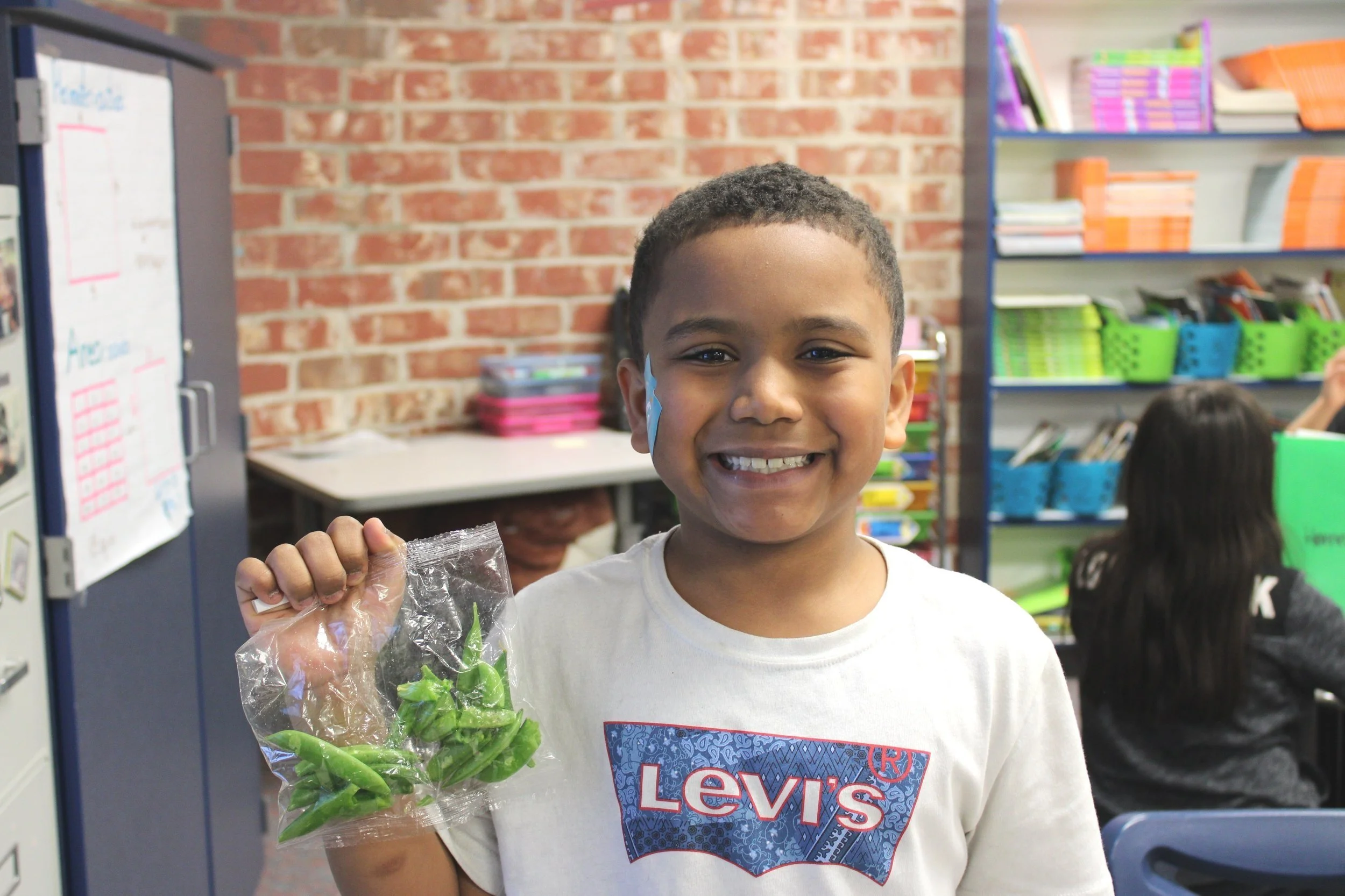 A young boy smiling and holding a plastic bag of green leafy vegetables in a classroom setting. He is wearing a white Levi's T-shirt with a design and a face sticker on his cheek. In the background, there are shelves filled with books and other classroom supplies, and a brick wall.