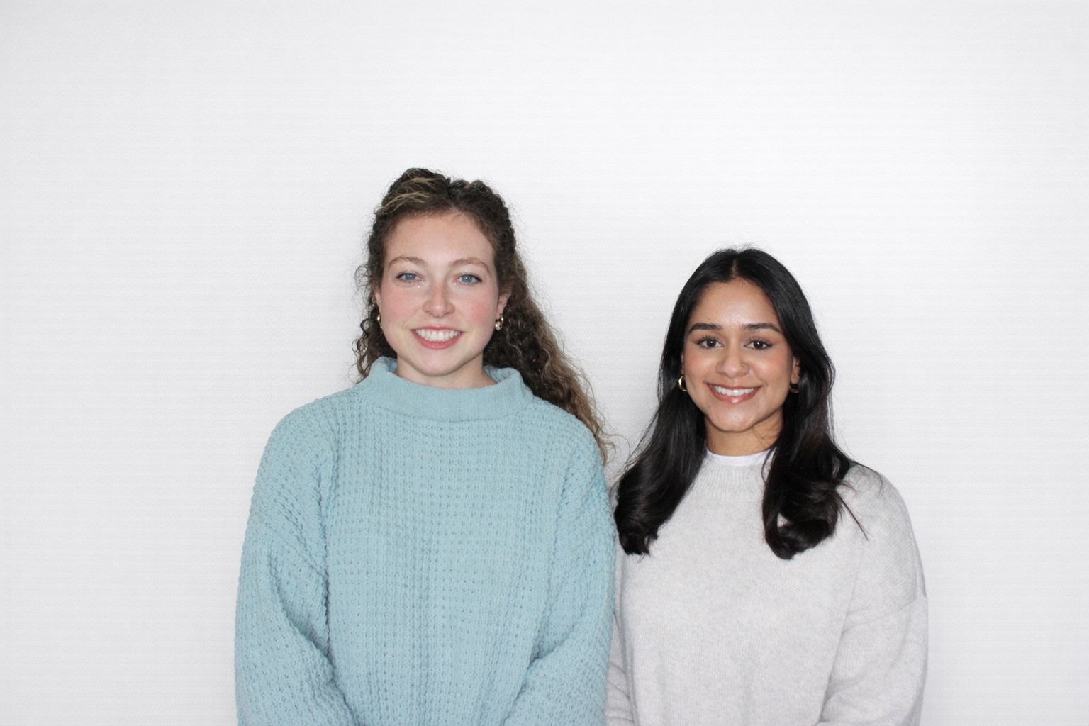 Two smiling women standing side by side against a plain white background, one with curly brown hair in a blue sweater, the other with straight black hair in a white sweater.