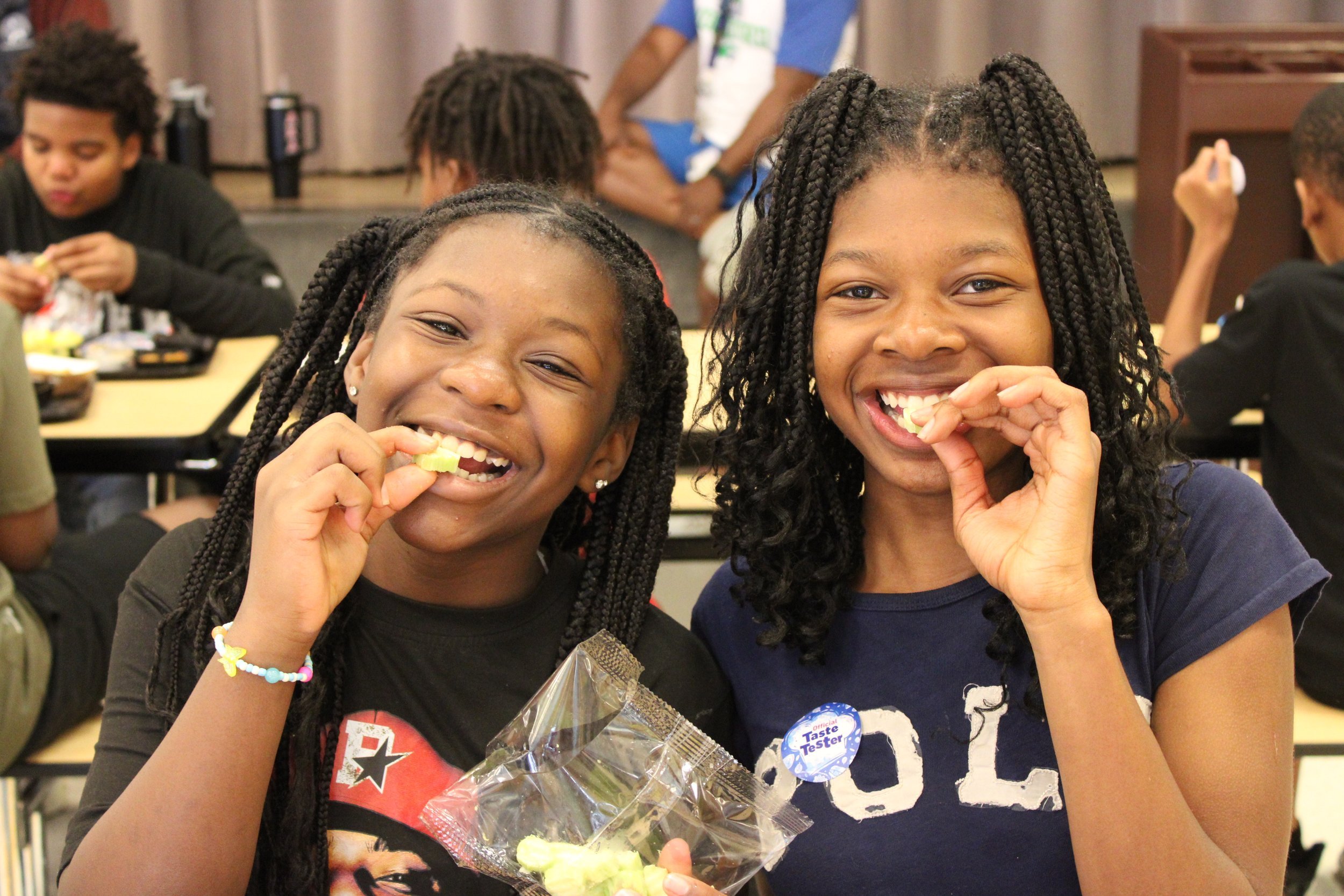 Two young girls with braided hair smiling and eating candy at a classroom or group setting, with other children in the background.