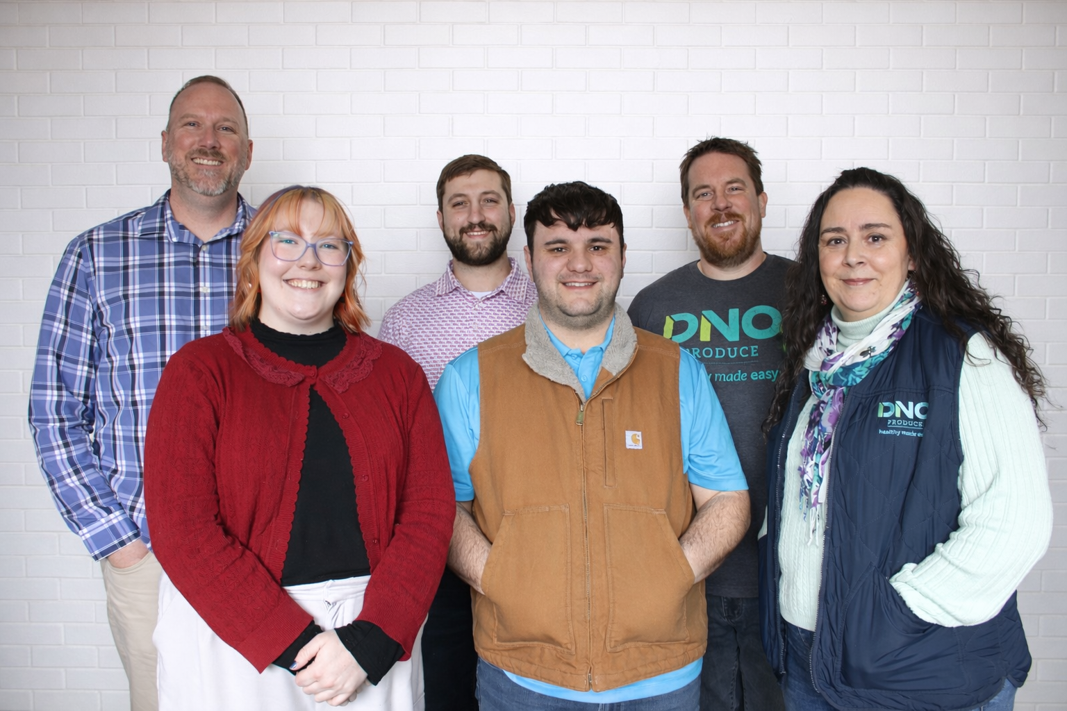 Group of six diverse adults standing against a white brick wall, smiling at the camera.