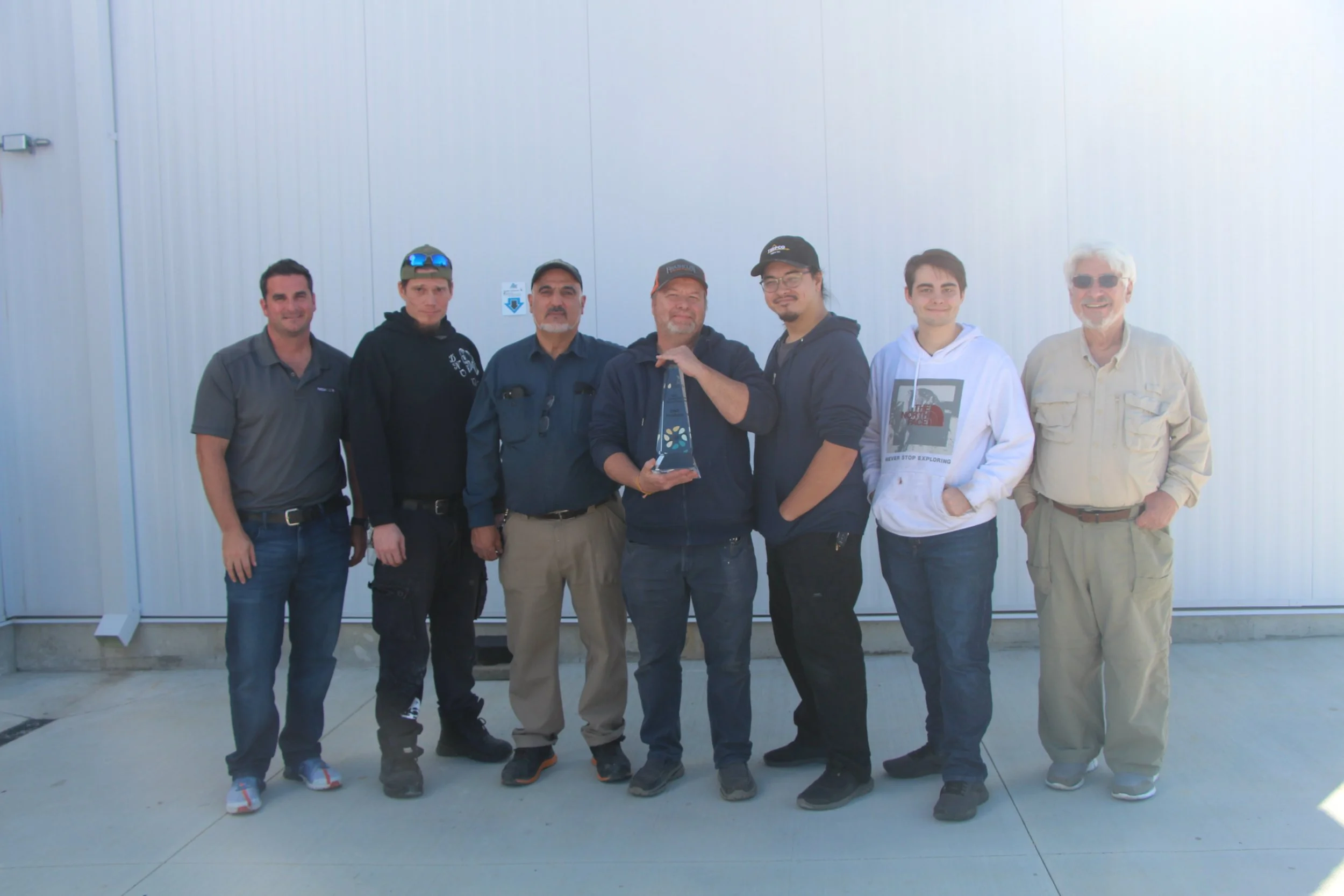 Group of seven men standing in front of a plain white wall, one holding a triangular award, everything in casual attire.