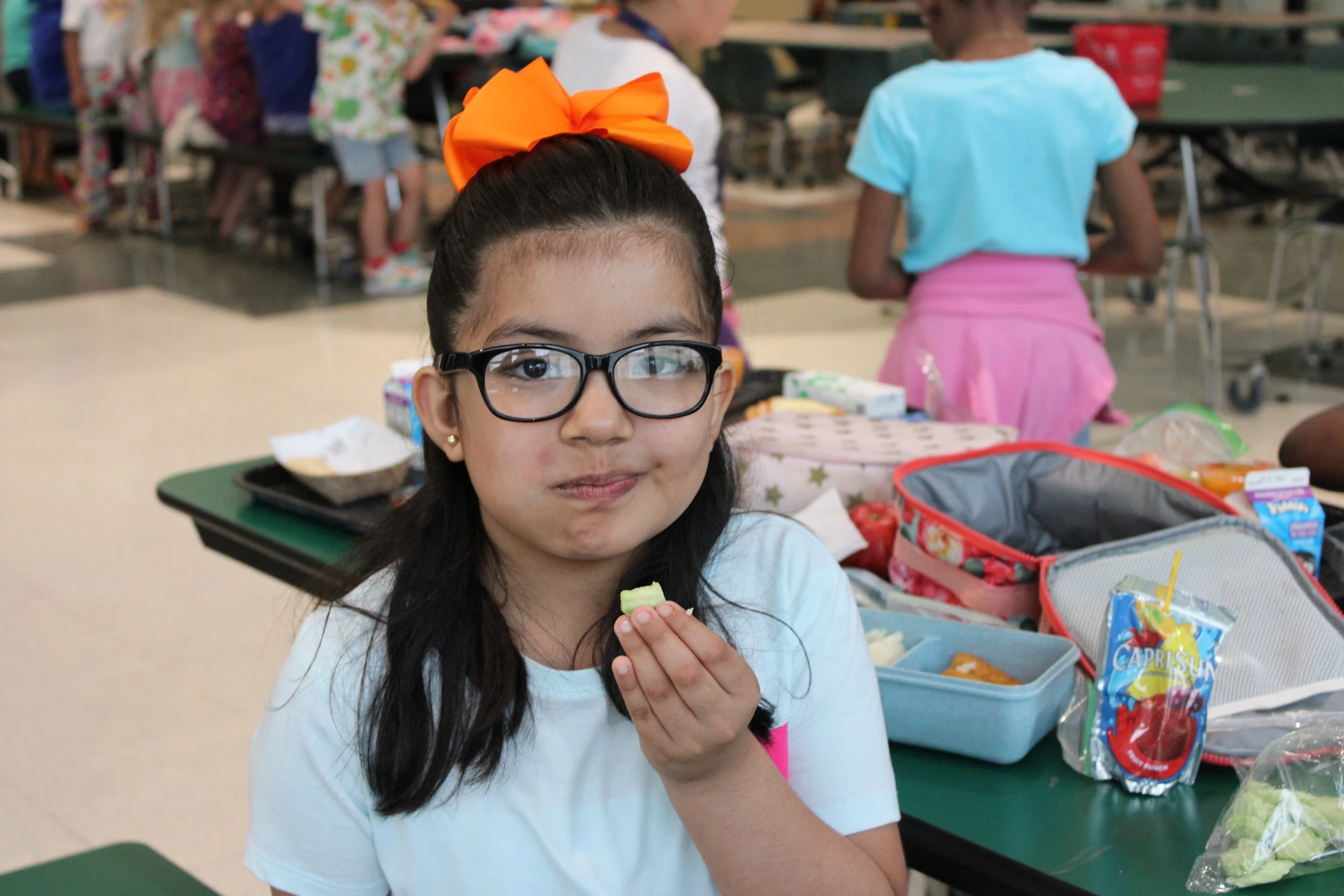 Young girl with glasses and an orange bow on her head, eating a piece of celery, sitting at a table in a cafeteria with lunch bags and food in front of her.