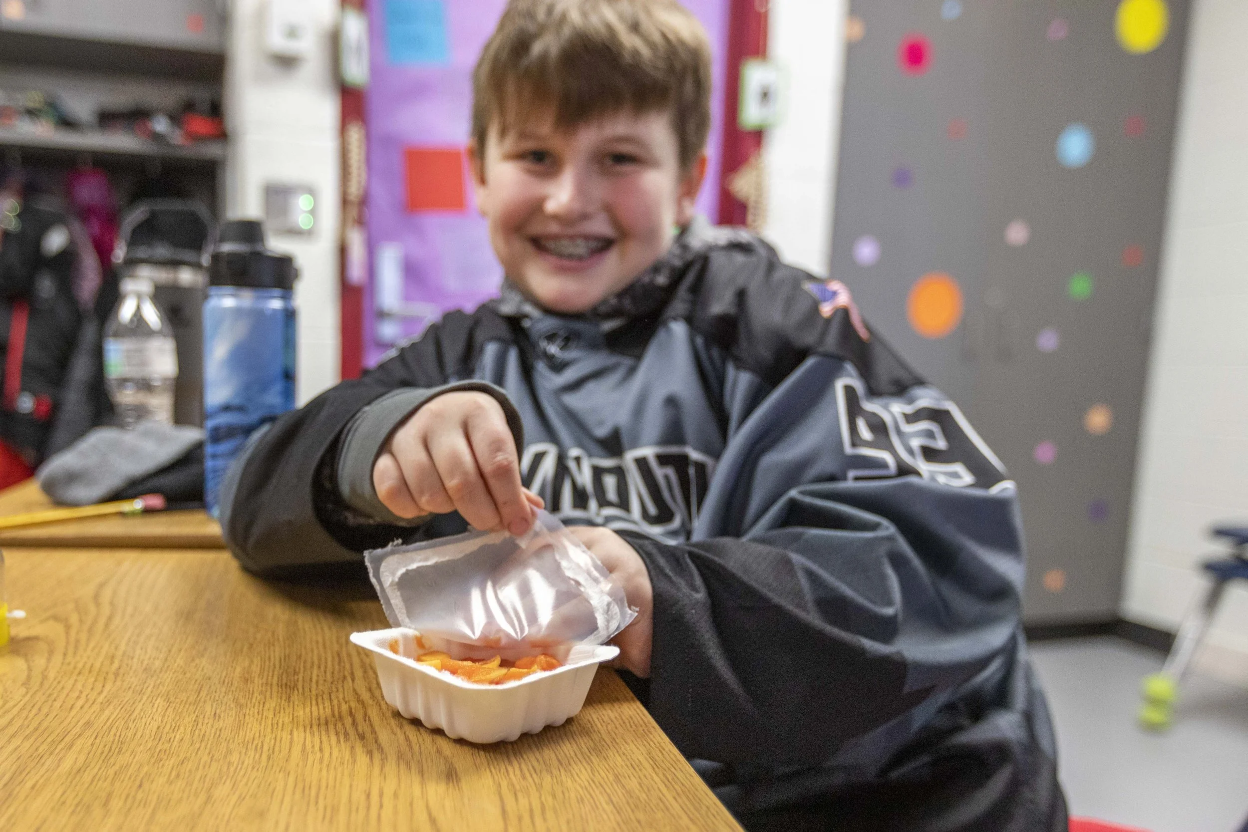 A young boy with braces smiling at the camera, sitting at a school table with a container of sliced carrots and a spoon in front of him, in a classroom with colorful decorations on the walls.