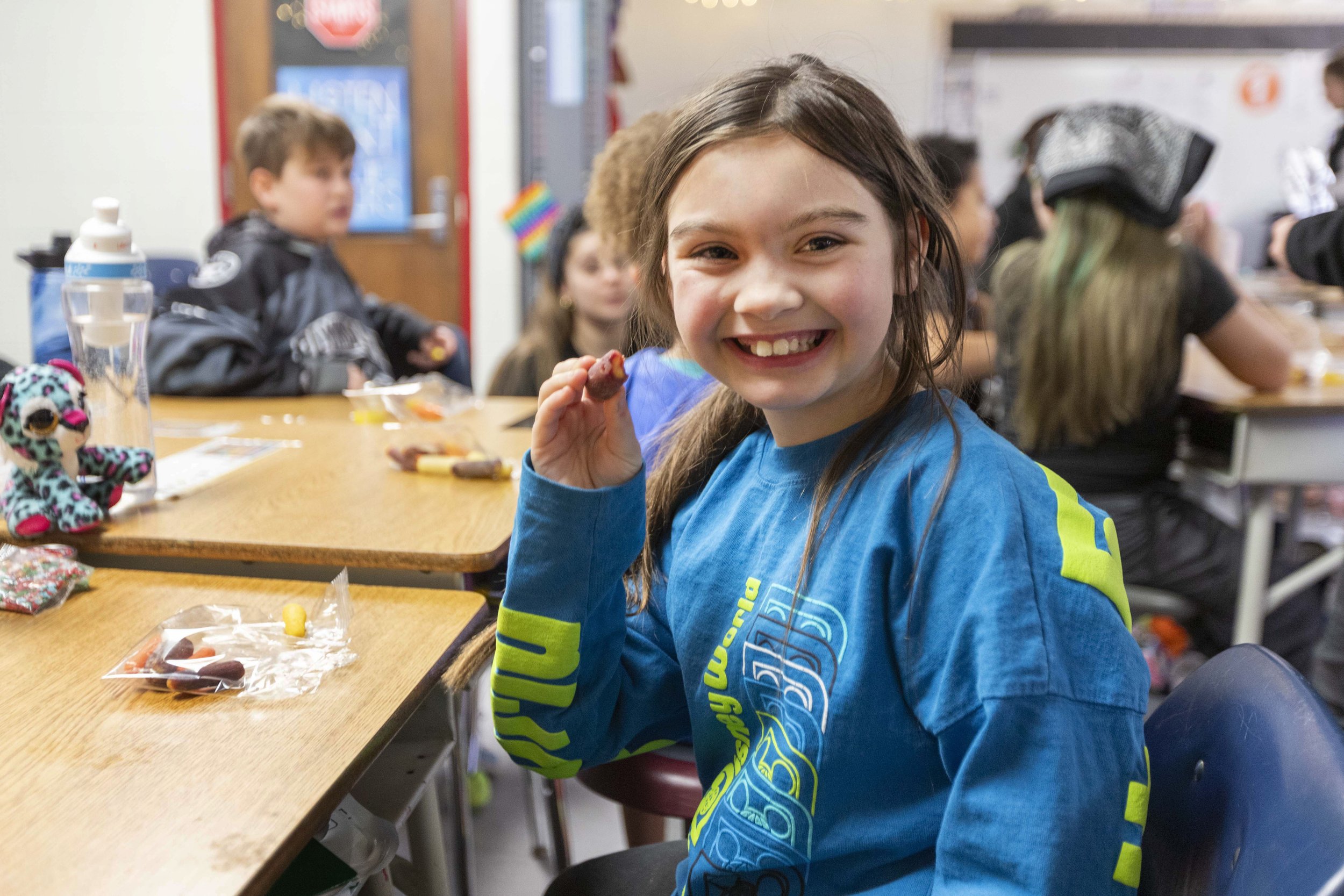 A young girl with long brown hair and a big smile, holding a cookie, sitting at a table with other children in a classroom setting.