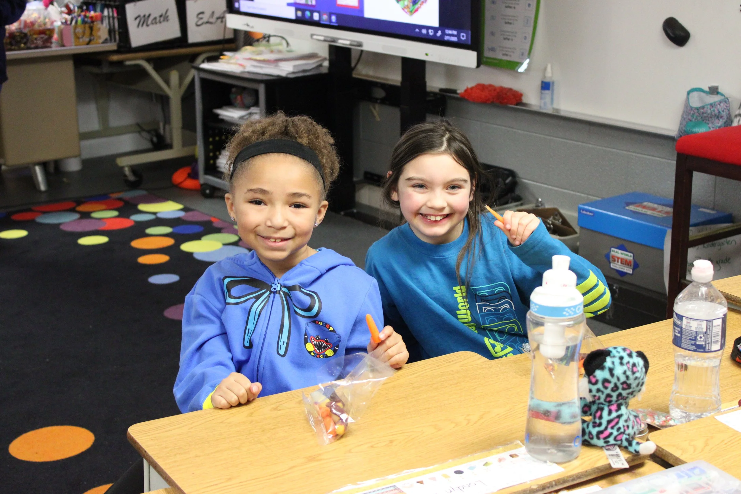 Two young girls smiling and sitting at a table in a classroom, surrounded by art supplies, drinks, and colorful decorations.