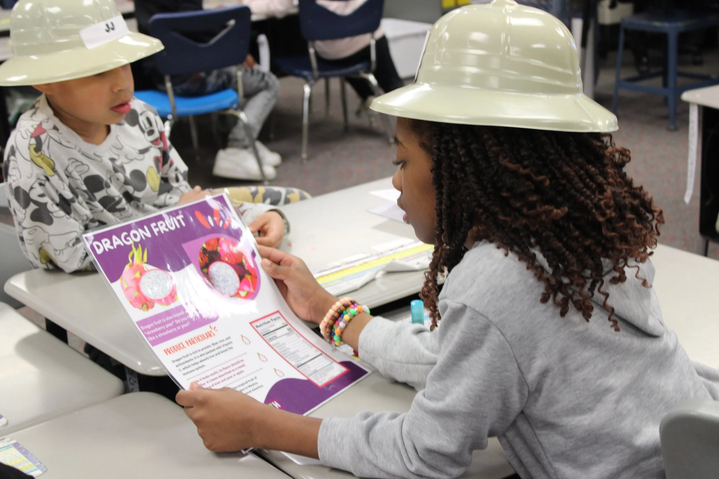 Two children wearing white safety helmets and sitting at a table in a classroom. One child is looking down at a colorful informational sheet about dragon fruit, which she is holding with both hands. The room has blue chairs and other students in the background.