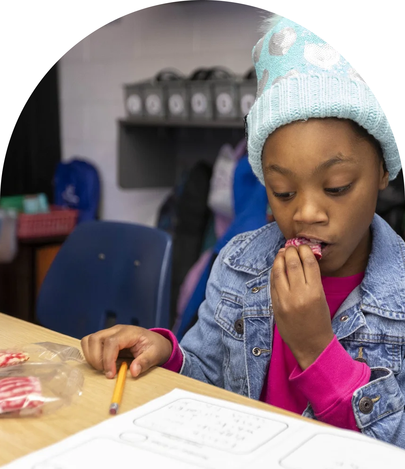 Young girl with a blue knit hat and pink long-sleeve shirt eating a cookie at a desk in a classroom.