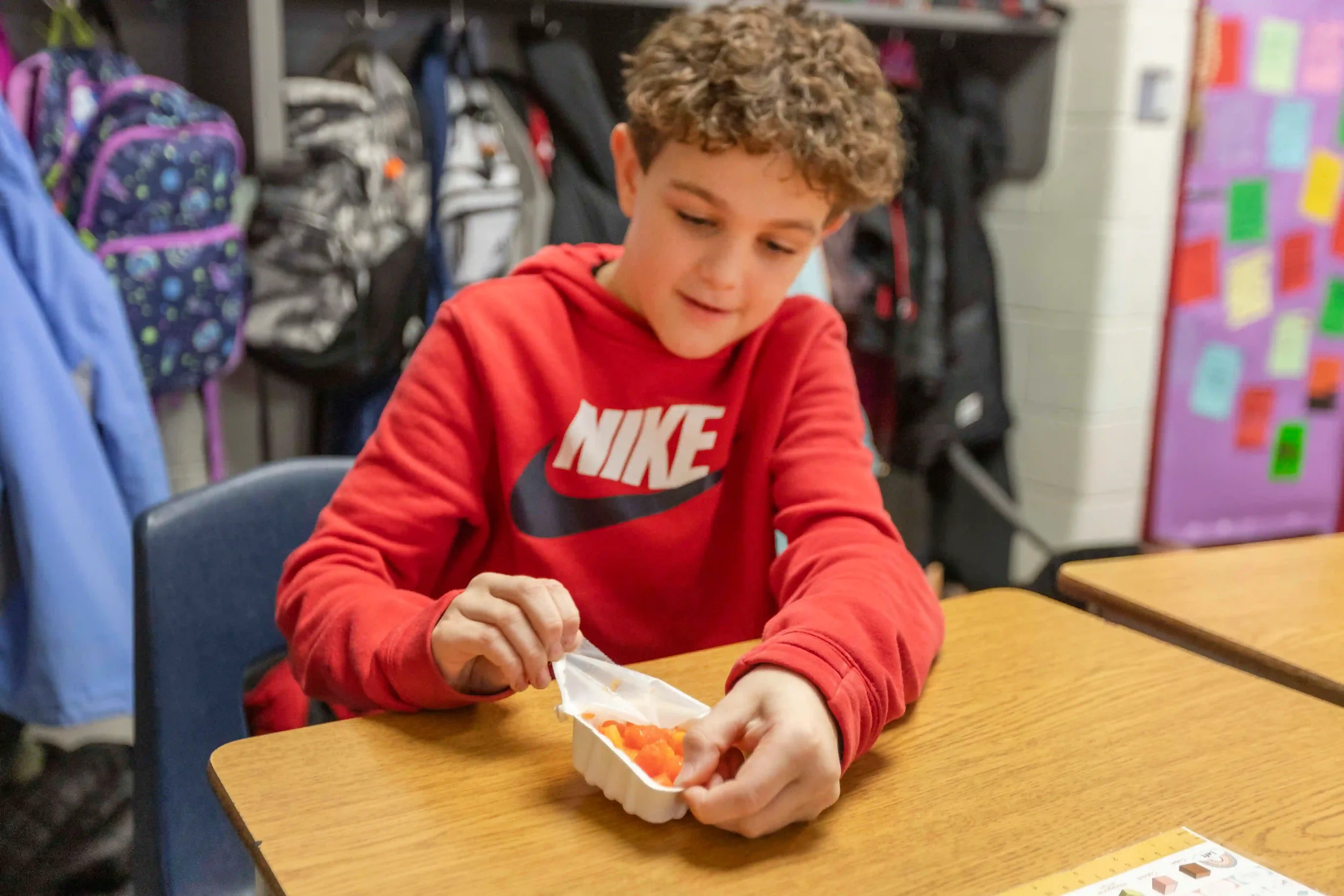 A young boy with curly hair wearing a red Nike sweatshirt sitting at a school desk opening a white container of orange-colored food.