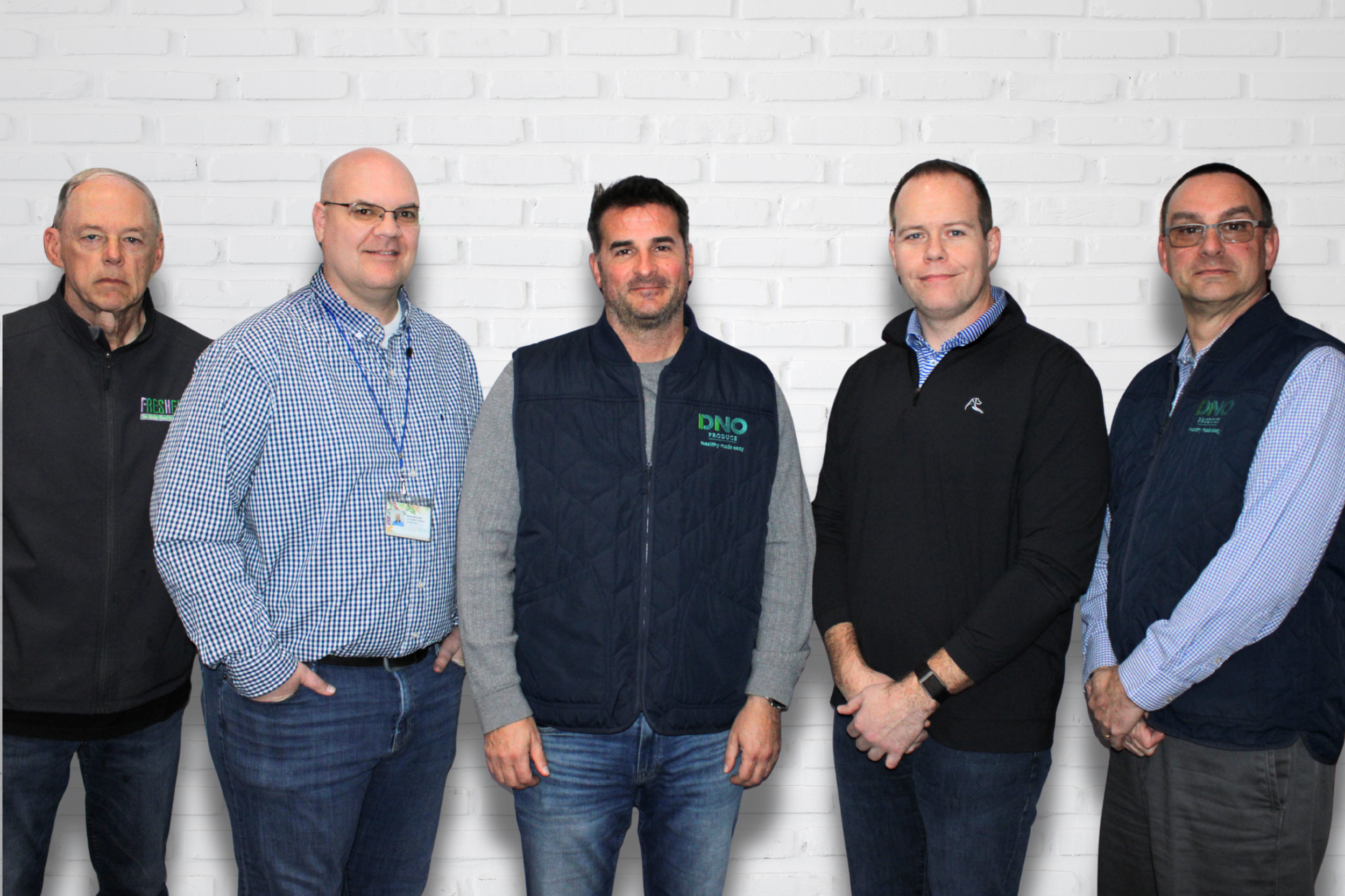 Group of five men standing against a white brick wall, dressed casually, with some wearing jackets and name tags.