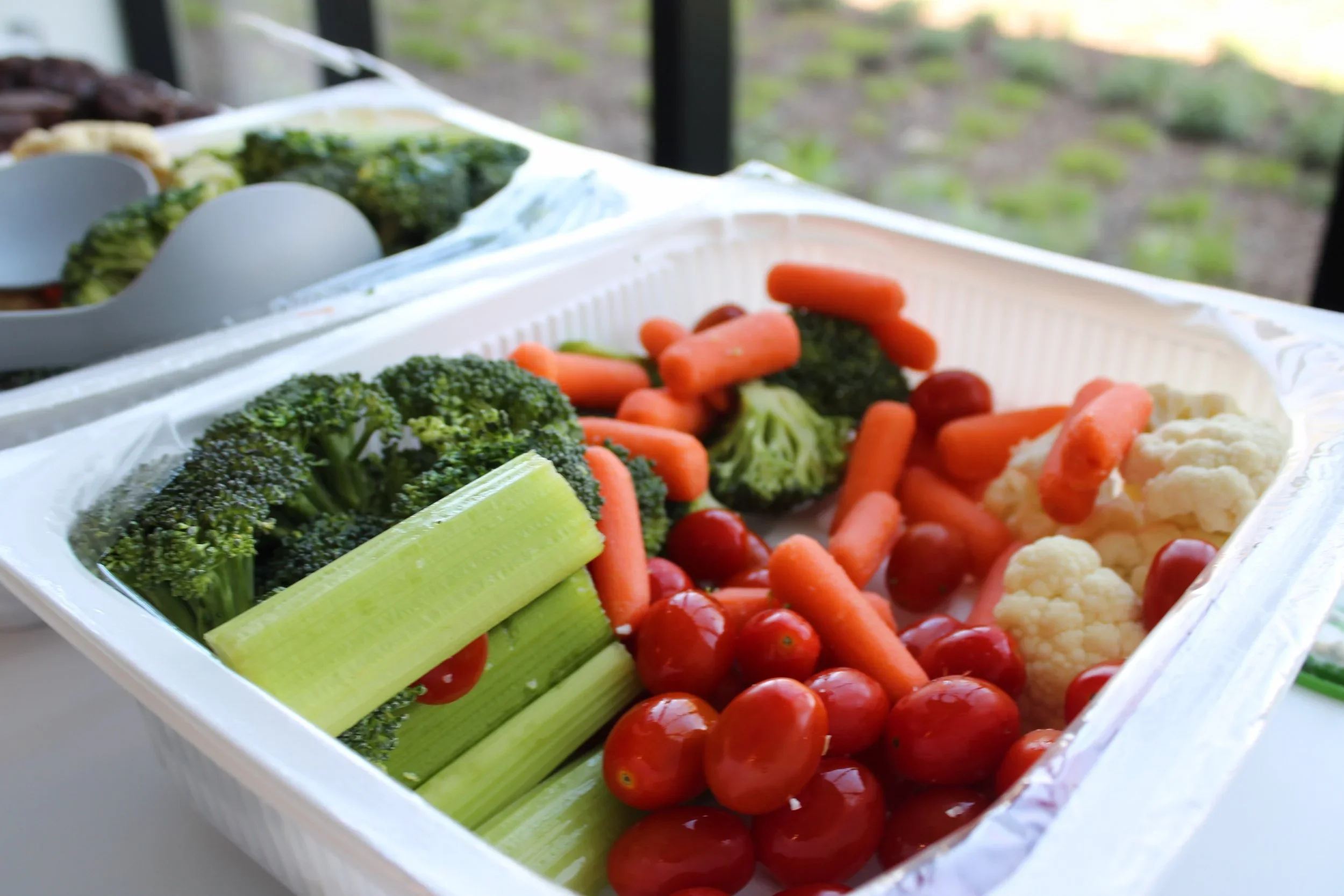 A white container filled with fresh vegetables, including cherry tomatoes, carrots, broccoli, cauliflower, celery, and spinach, with a blurred outdoor background.