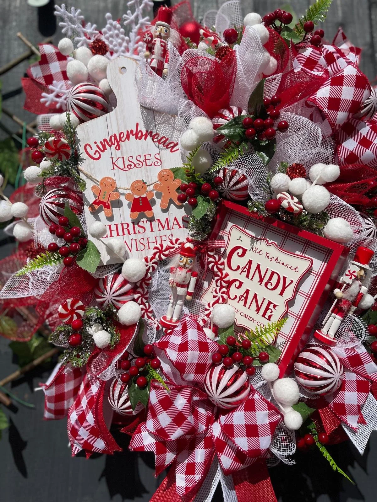 A Christmas wreath decorated with red and white ribbons, miniature gingerbread figures, candy canes, and fake snowballs. The wreath also features sprigs of holly with red berries and green leaves, a nutcracker figurine, and signs with holiday message