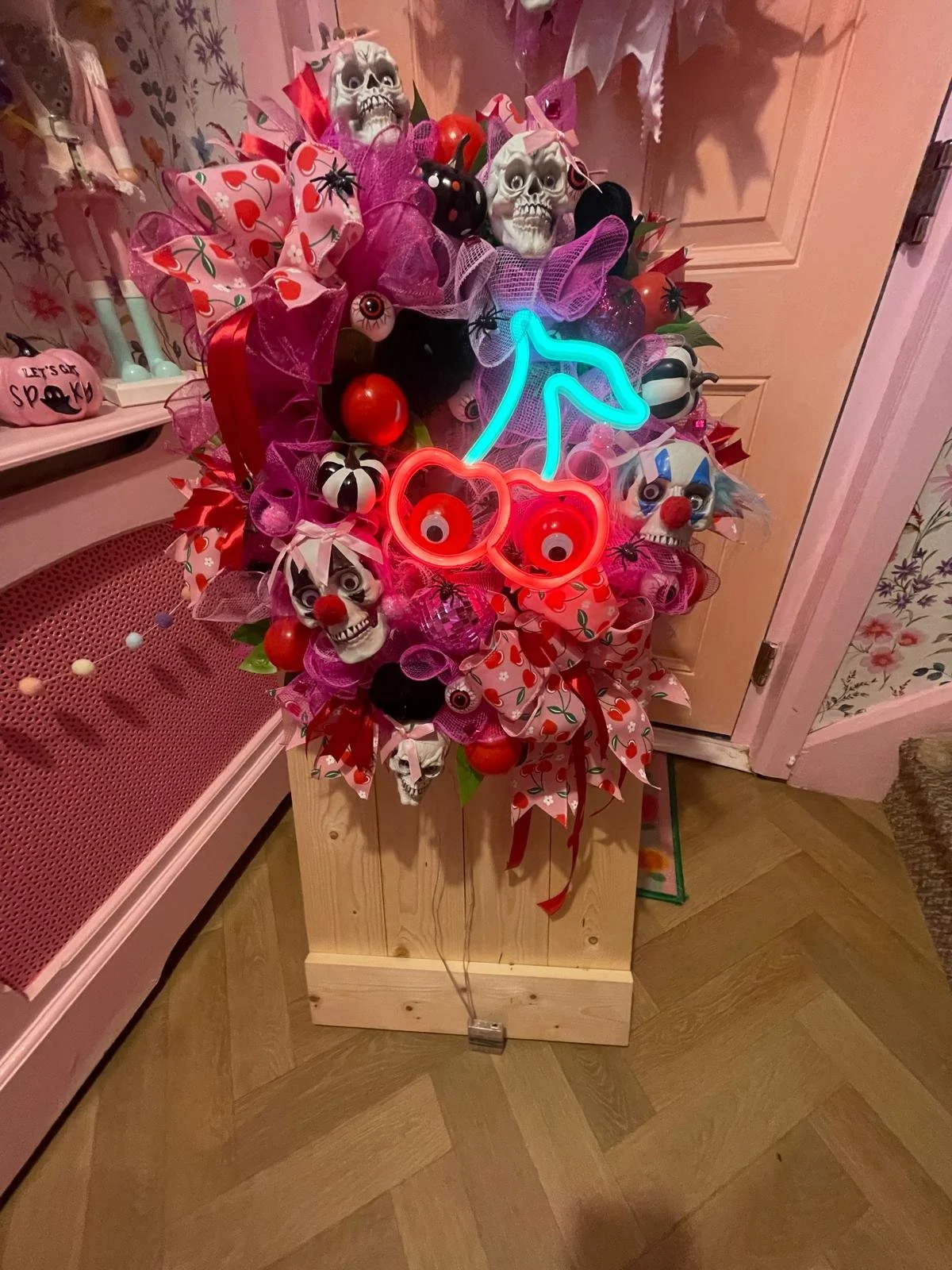 Decorative Halloween wreath featuring toy skulls, eyeballs, spiders, and neon lights shaped like a dinosaur, hearts, and glasses, with red, pink, and purple ribbons and bows.