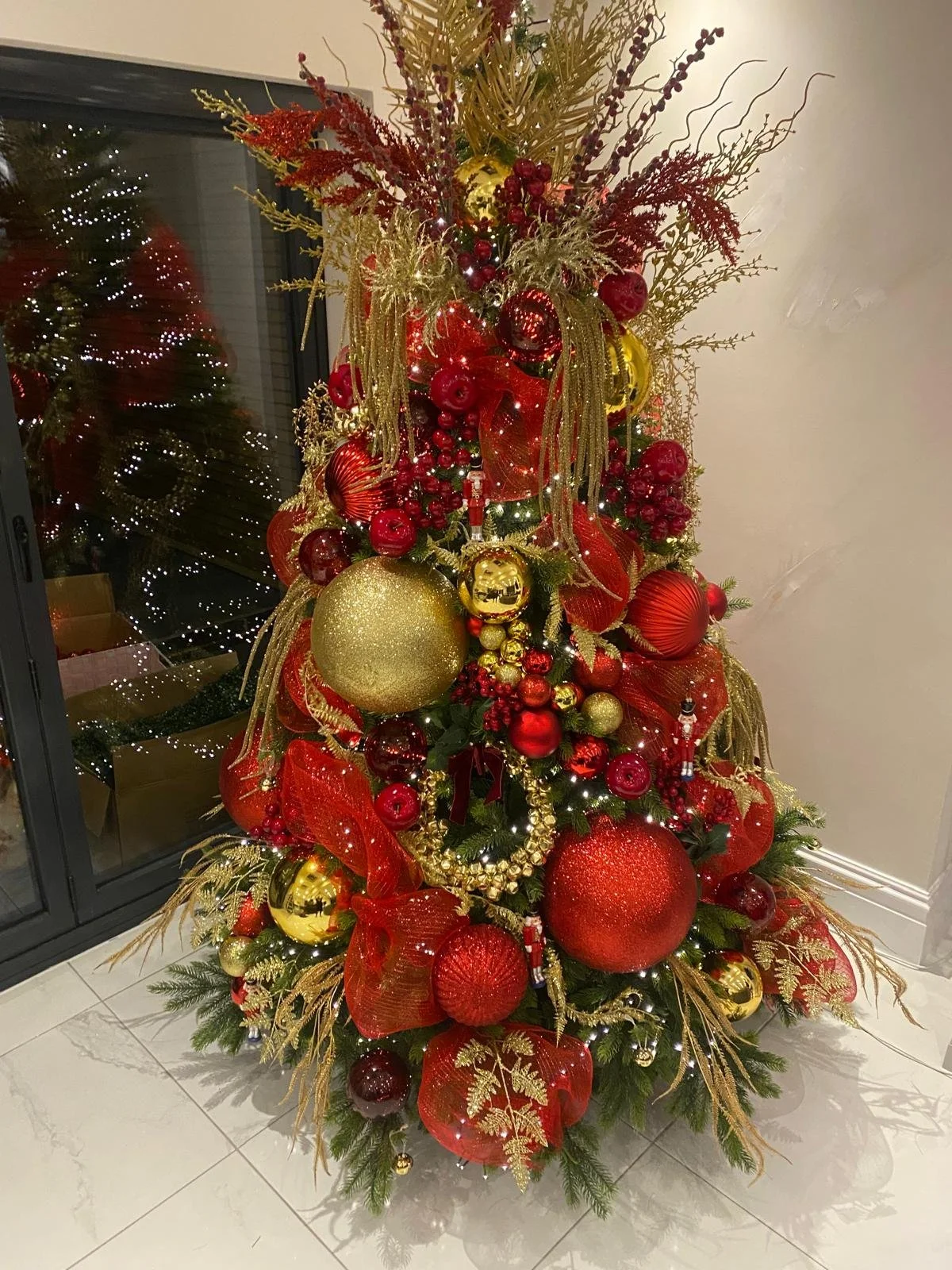 Decorated Christmas tree with red and gold ornaments, ribbons, and pine branches, standing on a tiled floor near a window.