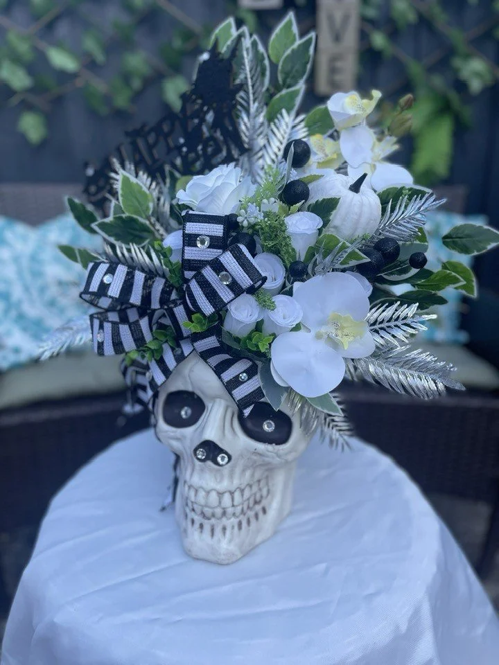 A decorative skull-shaped vase with a black and white striped bow, filled with artificial white flowers, green leaves, black berries, and metallic silver accents, arranged on a round white table.