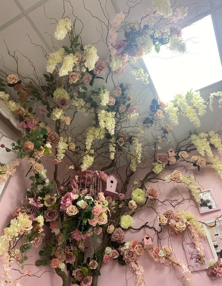 Decorative floral arrangement with pink, white, and cream roses, white hydrangeas, and branches, set against a pink wall with framed pictures and pink birdhouses.