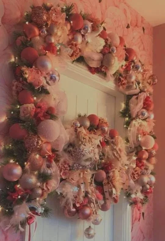 A Christmas wreath decorated with pink, red, and silver ornaments, ribbons, and feathers hanging on a door against pink floral patterned wallpaper.