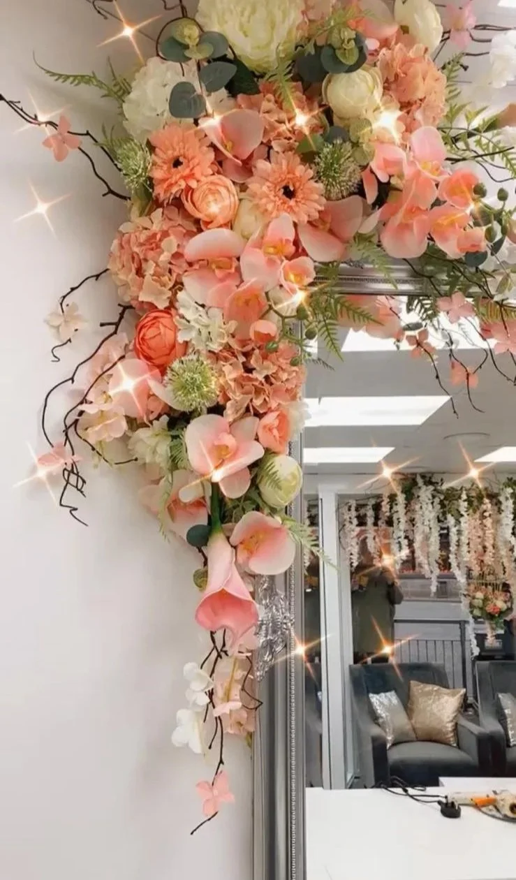 A floral arrangement with pink, peach, and white flowers, including roses and orchids, decorated with greenery and twigs, mounted on a wall near a mirror, with a sparkly filter effect.