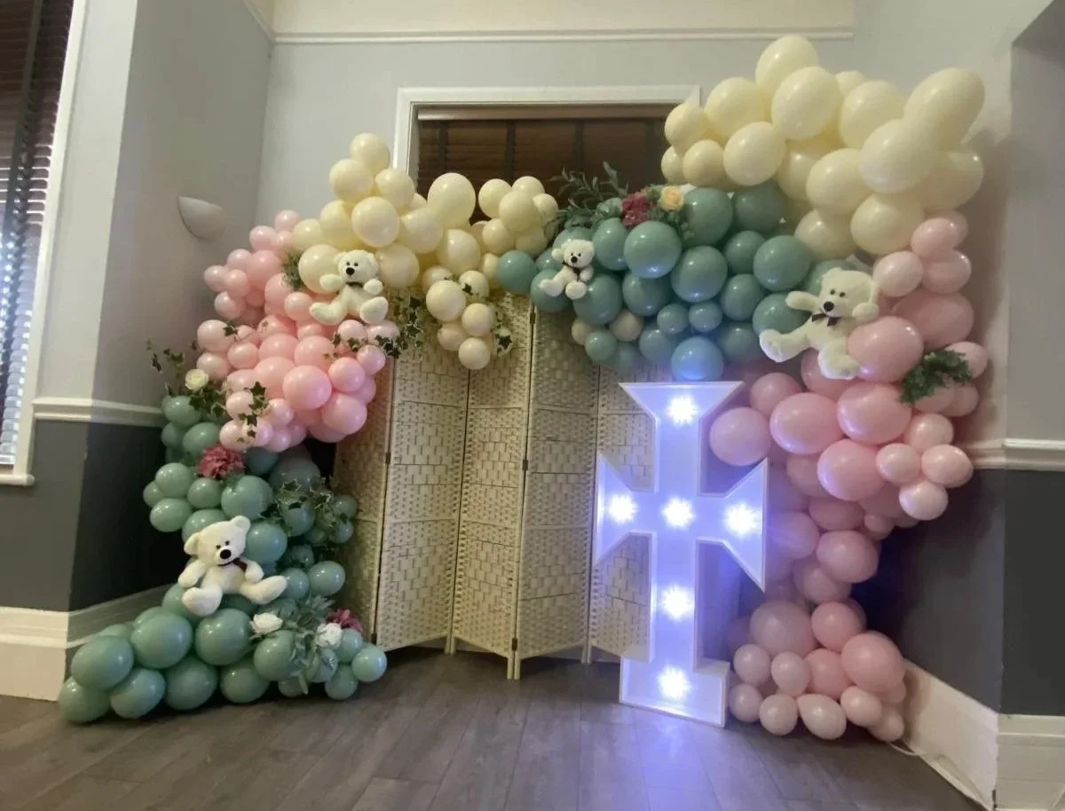 Decorative balloon arrangement with pastel pink, mint green, and cream balloons, teddy bears, flowers, a white illuminated star, and a letter "L" light at a party entrance.