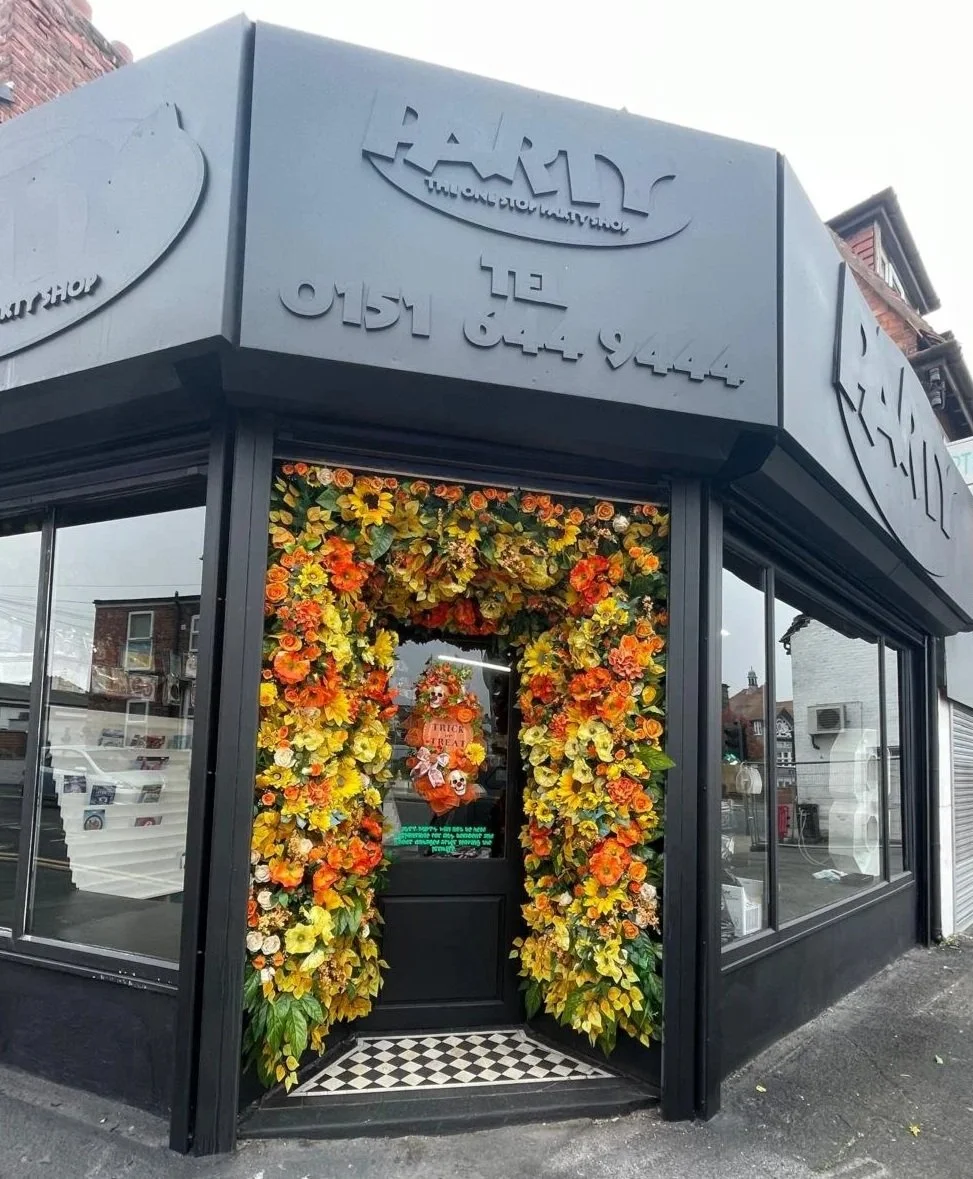 Storefront with black exterior, decorated with orange, yellow, and white flowers framing the black door, featuring a pumpkin decoration with a skull and 
