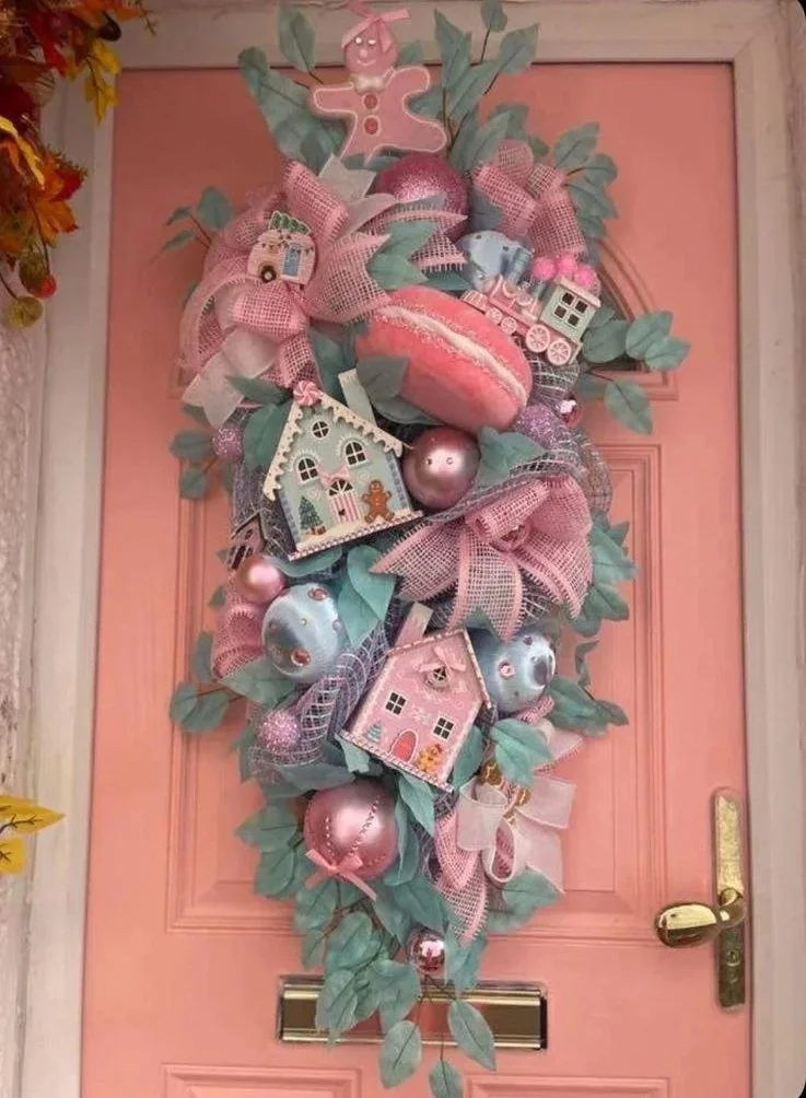 Decorative pink Christmas or holiday door wreath with pastel-colored ornaments, houses, a gingerbread figure, ribbons, and leaves arranged on a pink door.