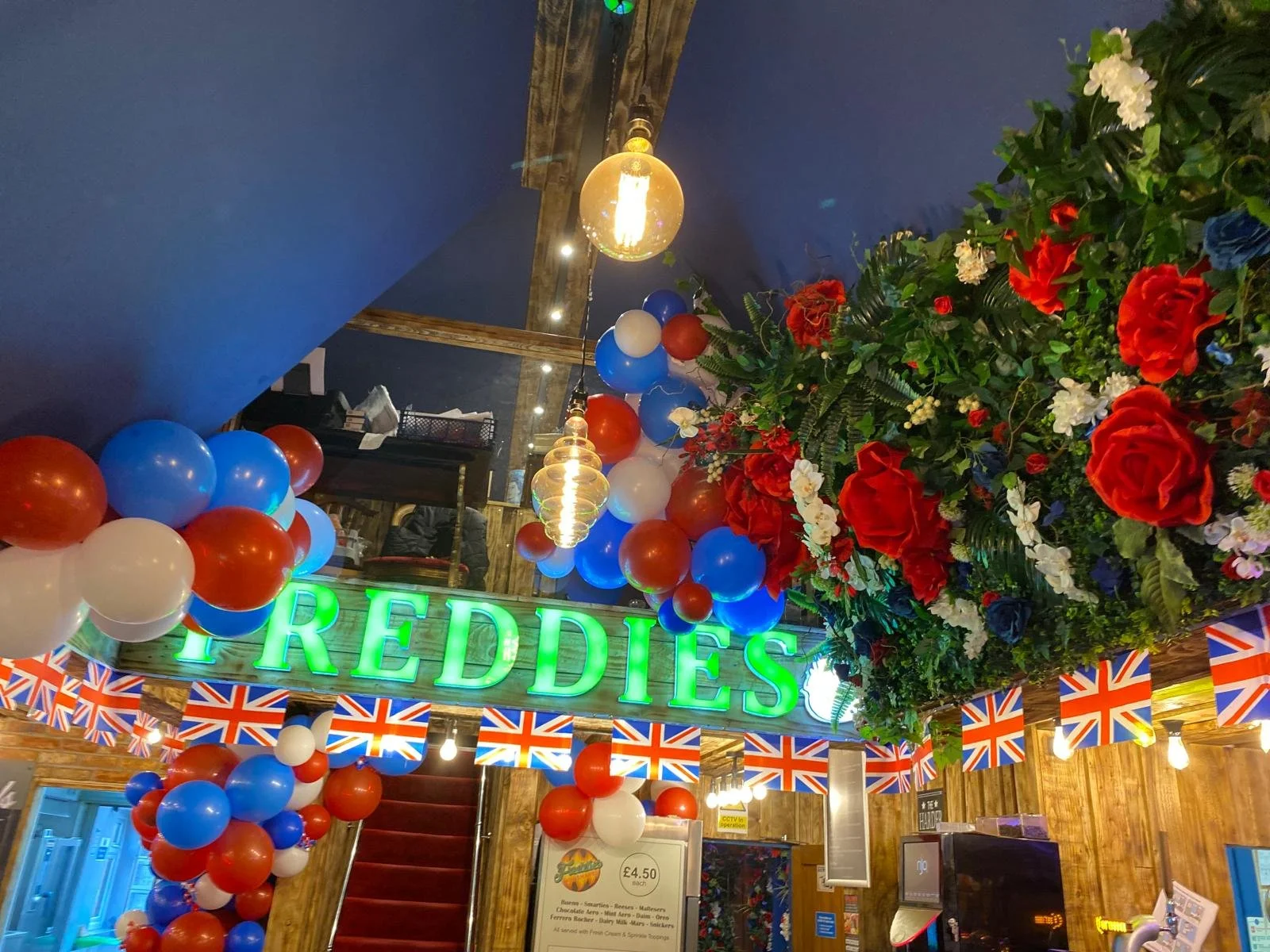 Decorated interior of a venue with balloons, string flags, a large floral arrangement, and a neon sign that reads 'REDDIES'.