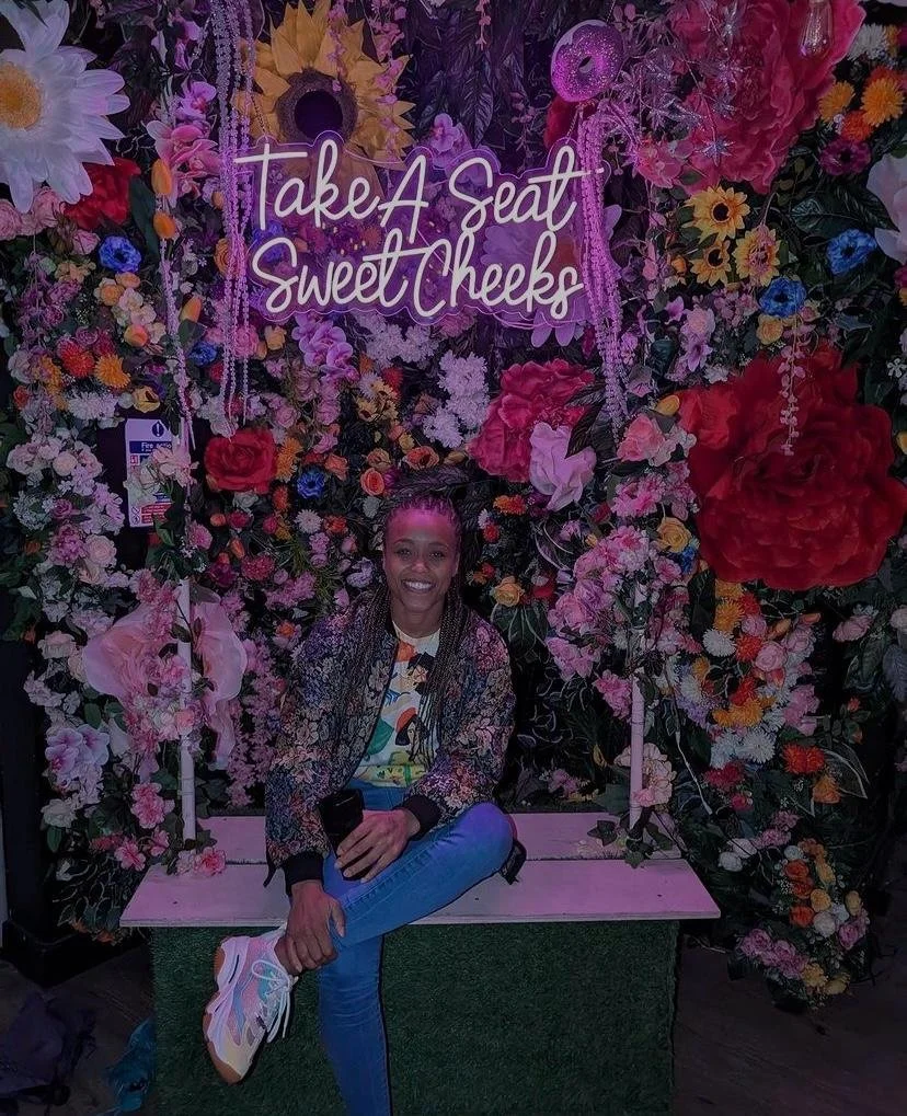 Woman sitting on a bench in front of a flower wall with a neon sign that says 'Take A Seat Sweet Cheeks,' surrounded by colorful flowers and decorations.