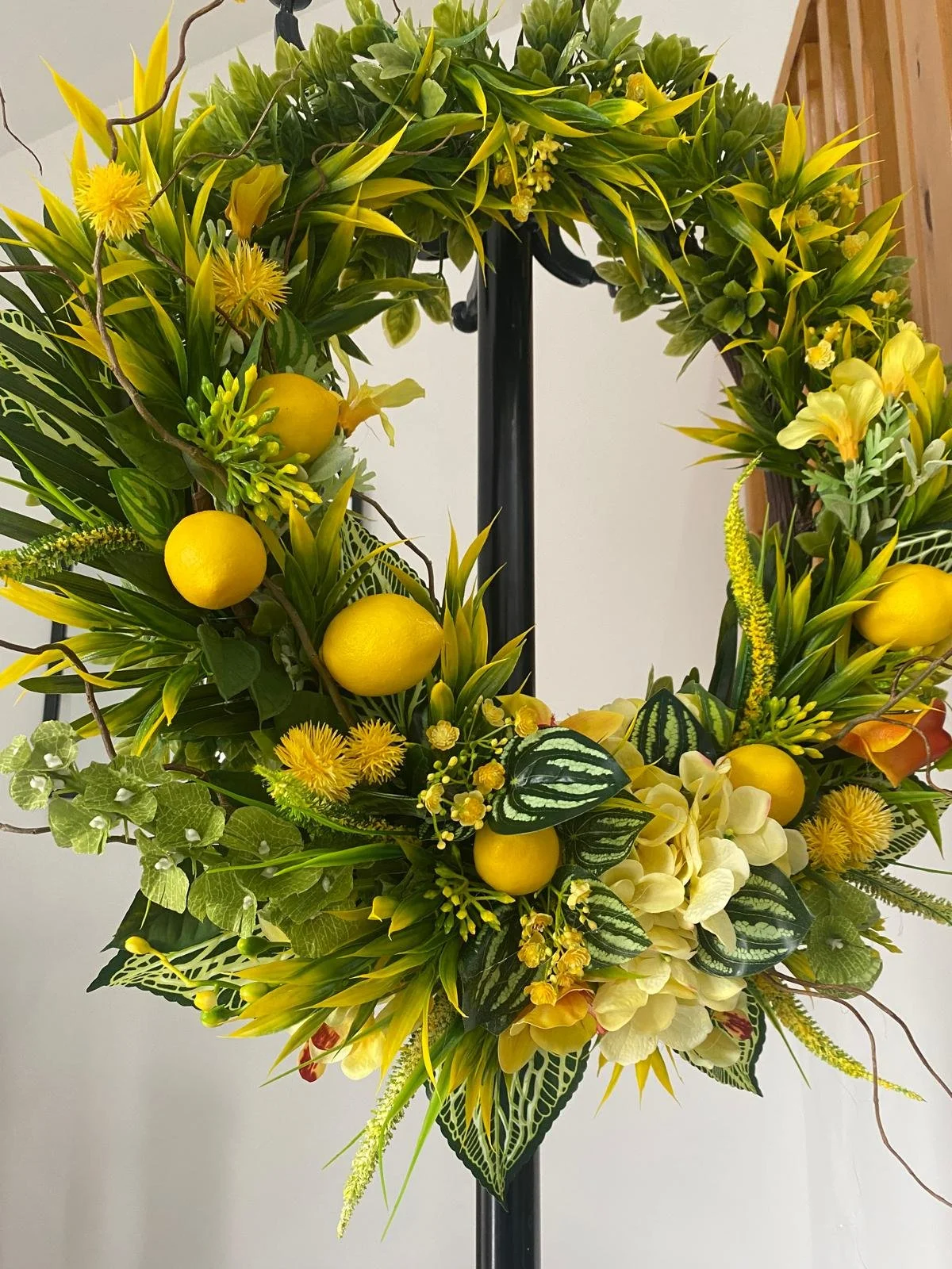 A floral arrangement in a wreath with yellow lemons, yellow flowers, green leaves, and various foliage on a black stand.
